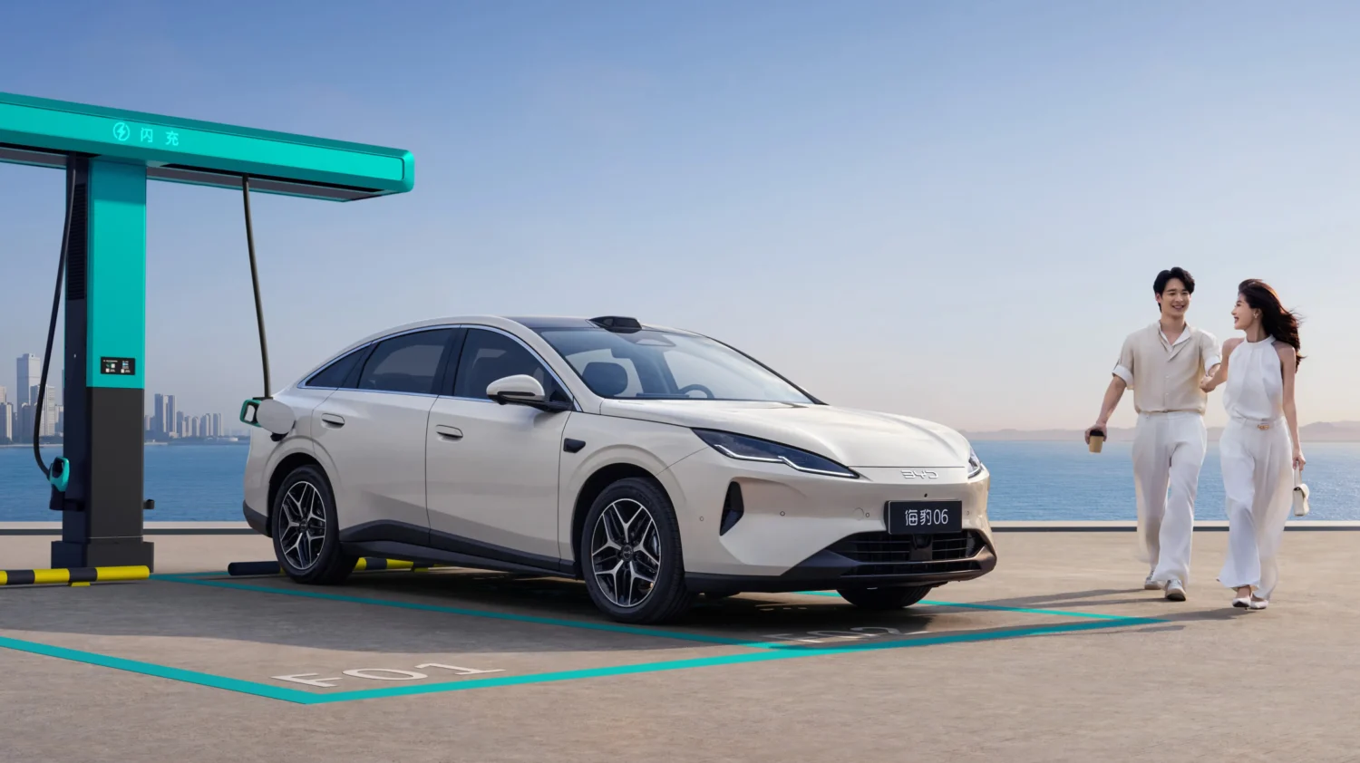 White electric sedan connected to a teal charging station by the water; a couple walks toward it, enjoying a sunny day.