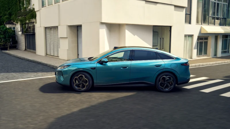 Teal blue modern sedan driving along a city street with white buildings in the background while crossing a crosswalk.