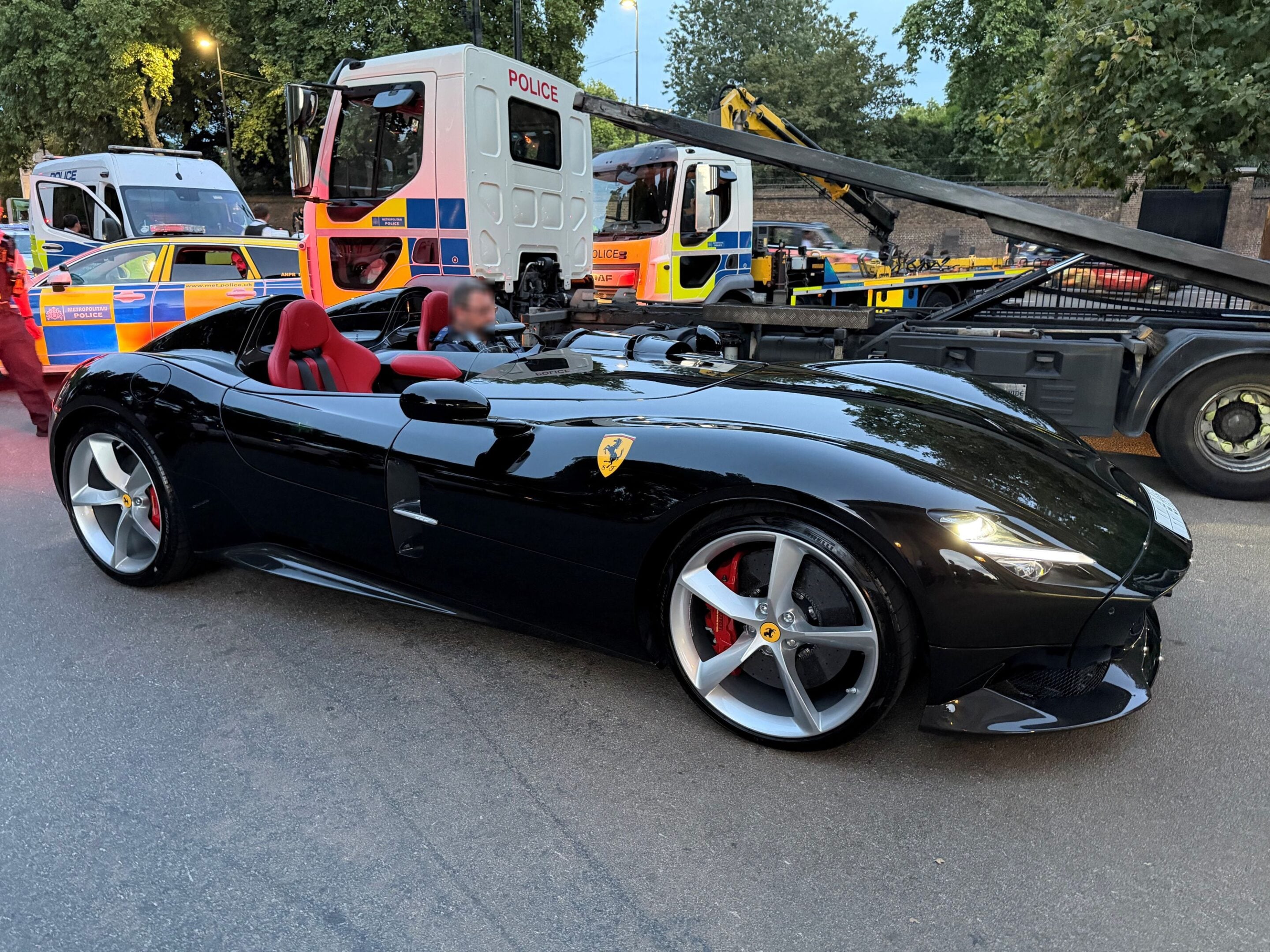 Black Ferrari convertible with red seats in the foreground, with police vans and a tow truck in the background at an emergency scene.