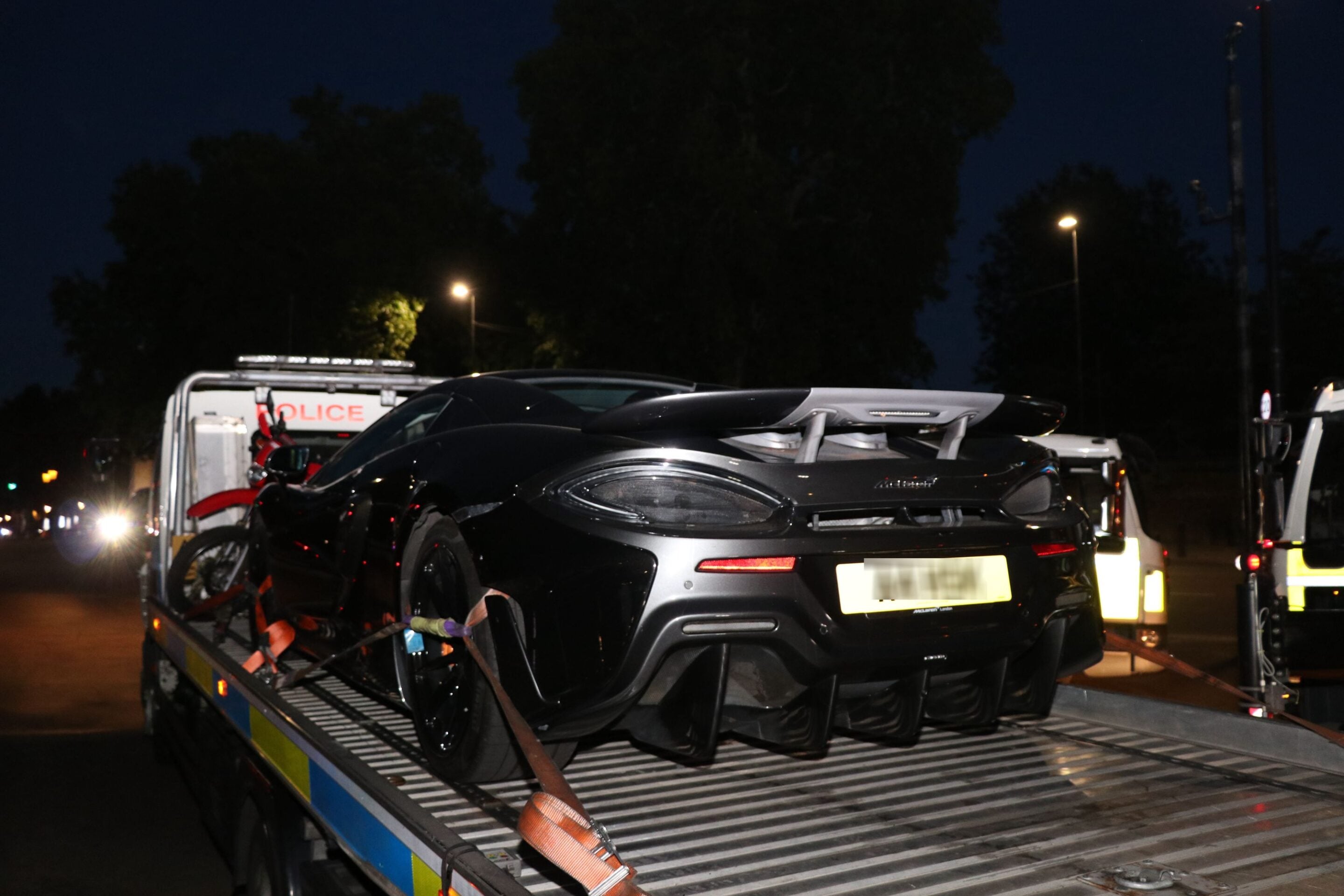 Black sports car strapped onto a tow-truck trailer at night, with other vehicles in the background and streetlights nearby.