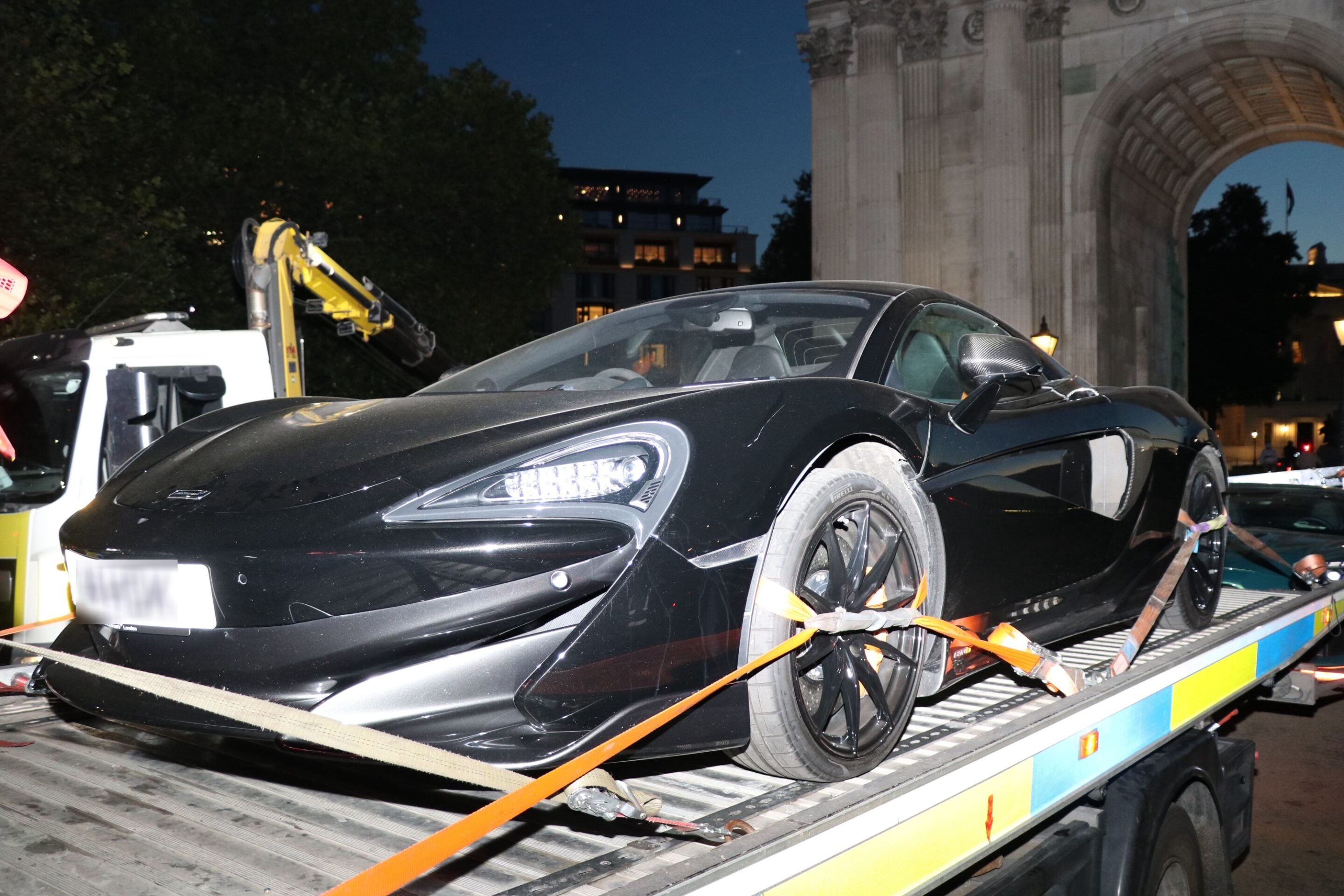 Black sports car secured on a flatbed tow truck at night, orange straps tightening it, with a stone arch in the background.