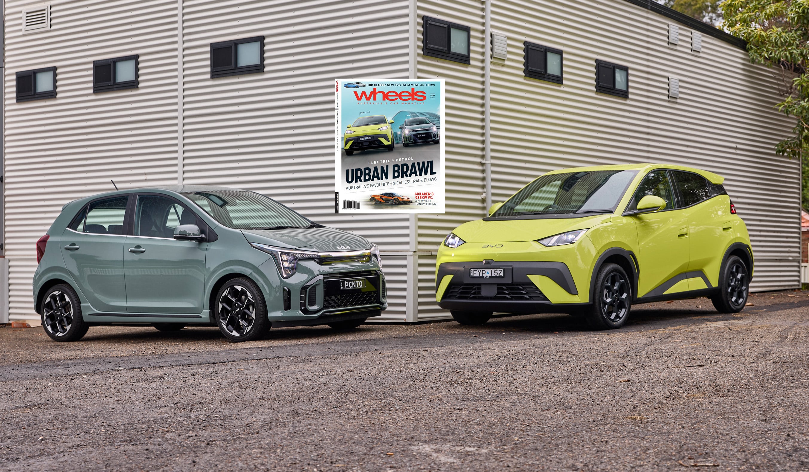Two modern cars parked side by side in front of a beige corrugated metal wall, with a Wheels magazine poster between them.