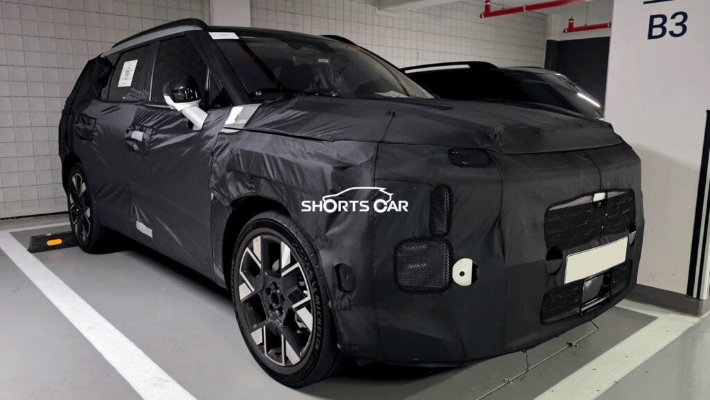 Camouflaged SUV prototype parked in a garage; black wrap conceals design, Shorts Car watermark visible.