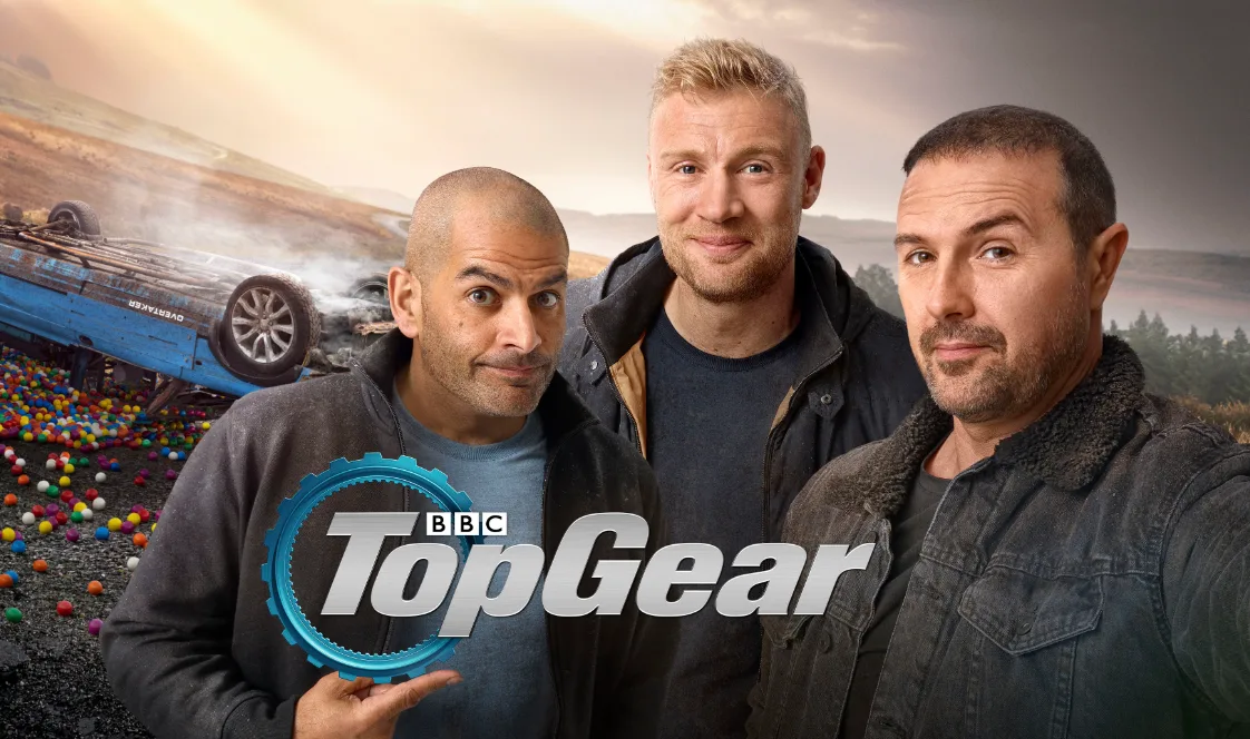 BBC Top Gear hosts pose together with an overturned blue car and a field of colorful balls in the background.