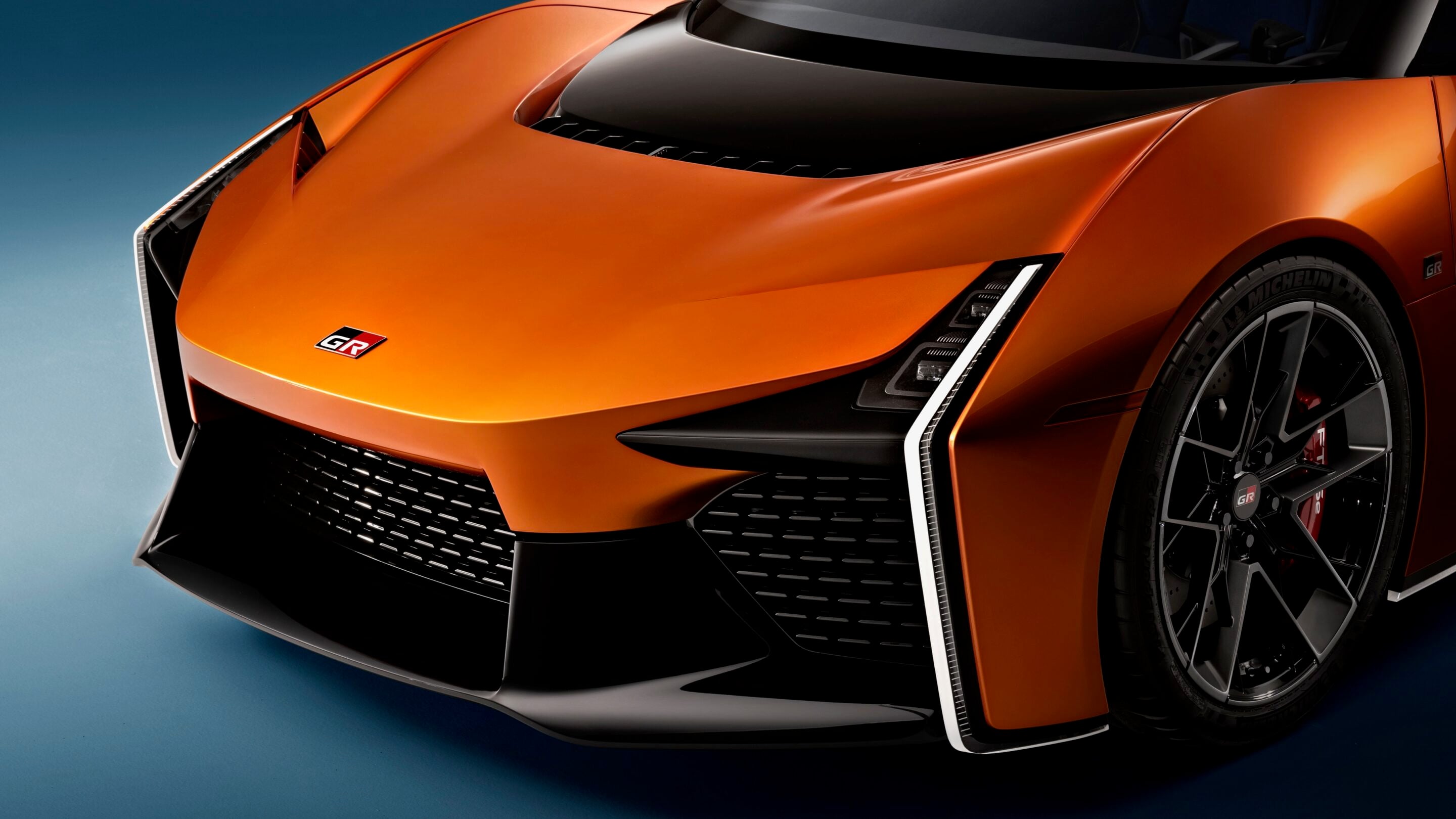 Close-up of an orange sports car's front end, showing the sleek hood, angled headlight cluster, and lower grille with a GR badge on the hood edge, set against a blue background.