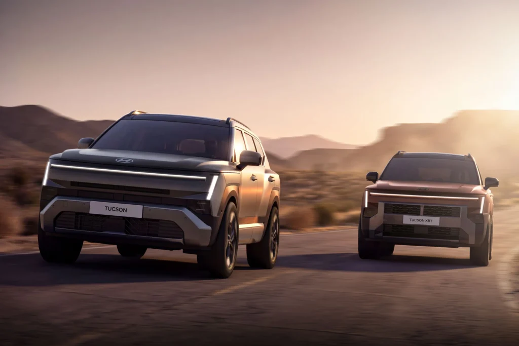 Two silver Hyundai SUVs (Tucson and Tucson XRT) drive side by side along a desert highway at sunset, with blurred mountains in the background and warm light reflecting off the vehicles.