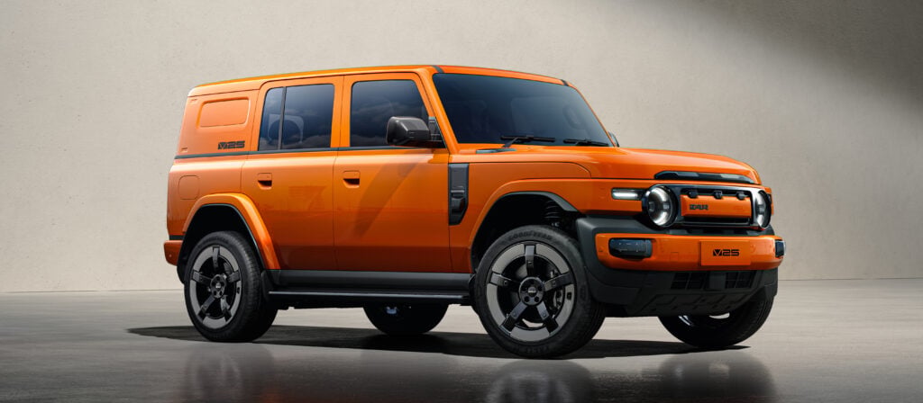 Orange off-road SUV shown in a three-quarter front view indoors with large black alloy wheels and rugged bumper, parked on a reflective floor.
