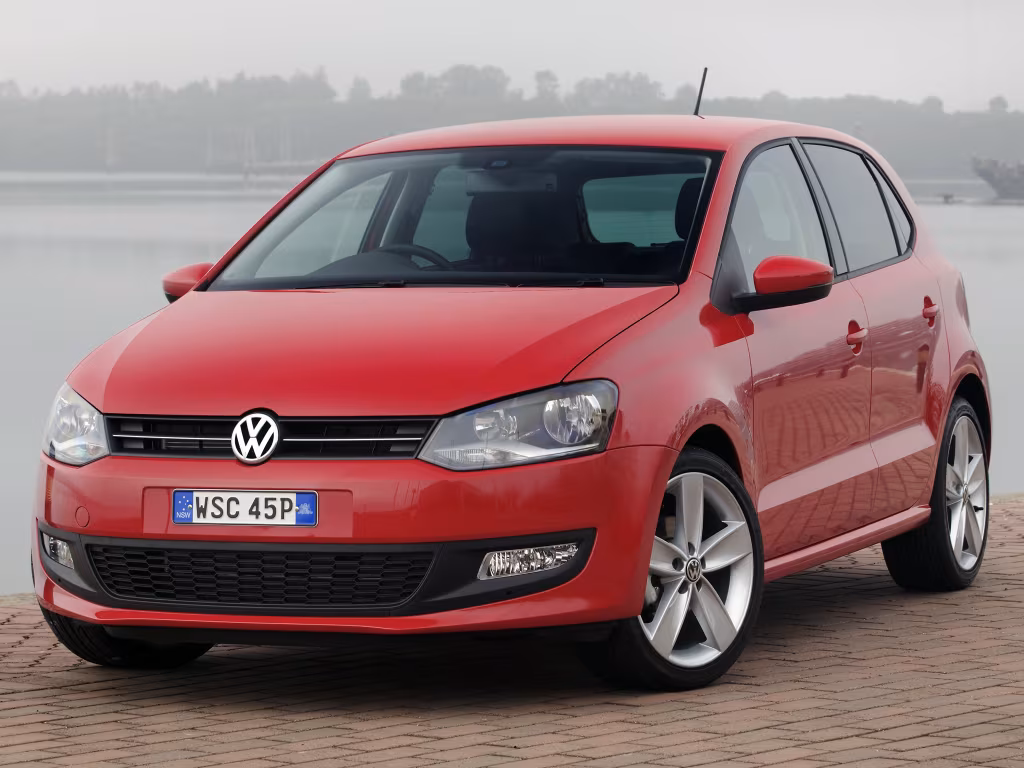 Red Volkswagen hatchback parked on a brick-paved waterfront area, showing the front and driver's side with a license plate visible.