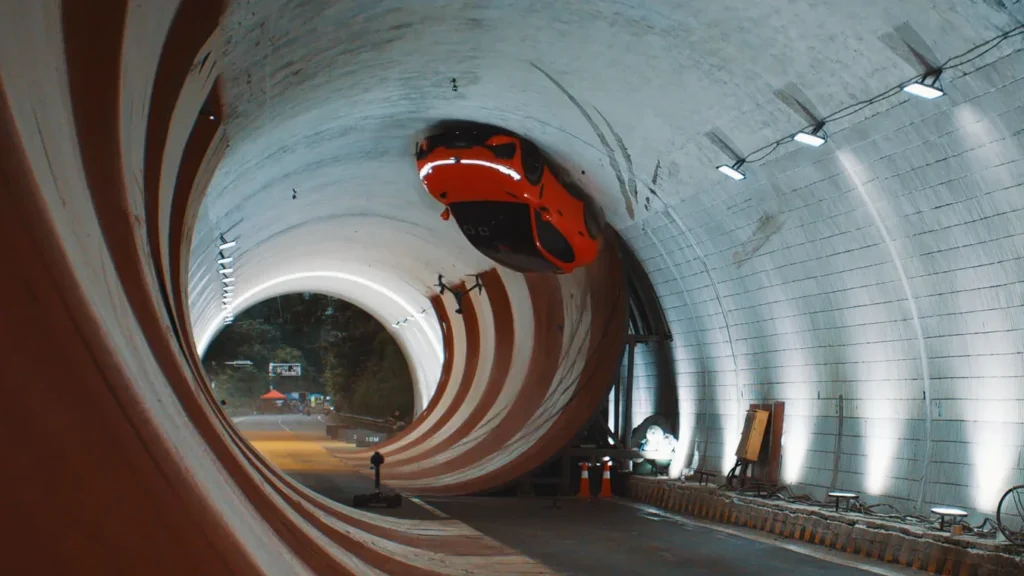 Orange sports car suspended inside a large circular test tunnel with curved white walls and construction equipment nearby.