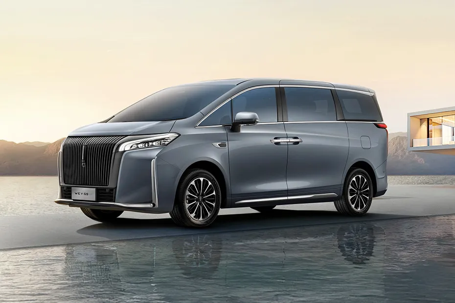 Gray luxury minivan shown from the front‑left, parked on a reflective surface at sunset with a bold vertical grille and chrome accents.