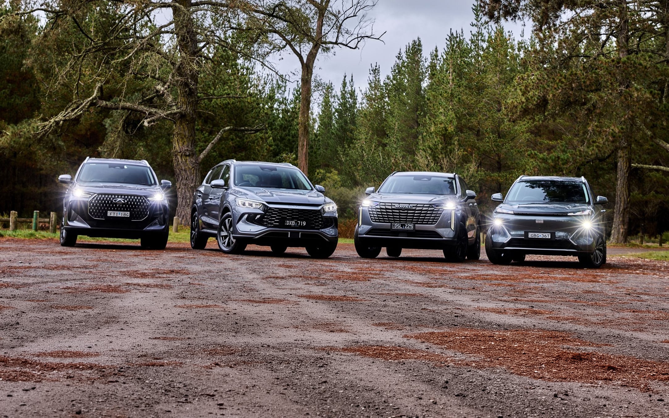 Four SUVs parked in a row on a gravel lot in a forested area, headlights on, trees in the background.