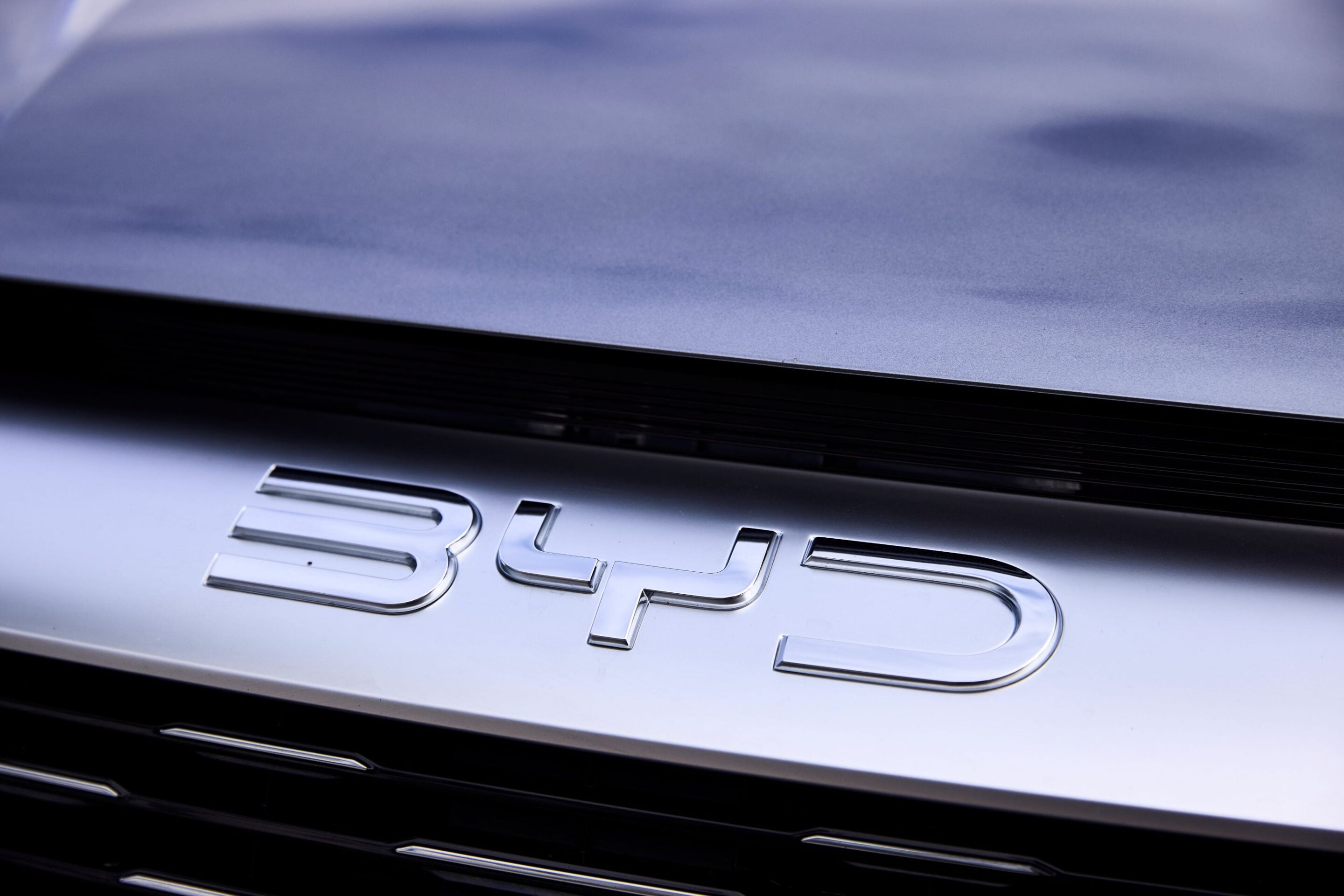 Close-up of a shiny chrome car badge on a silver trunk, with blurred dark grille below.