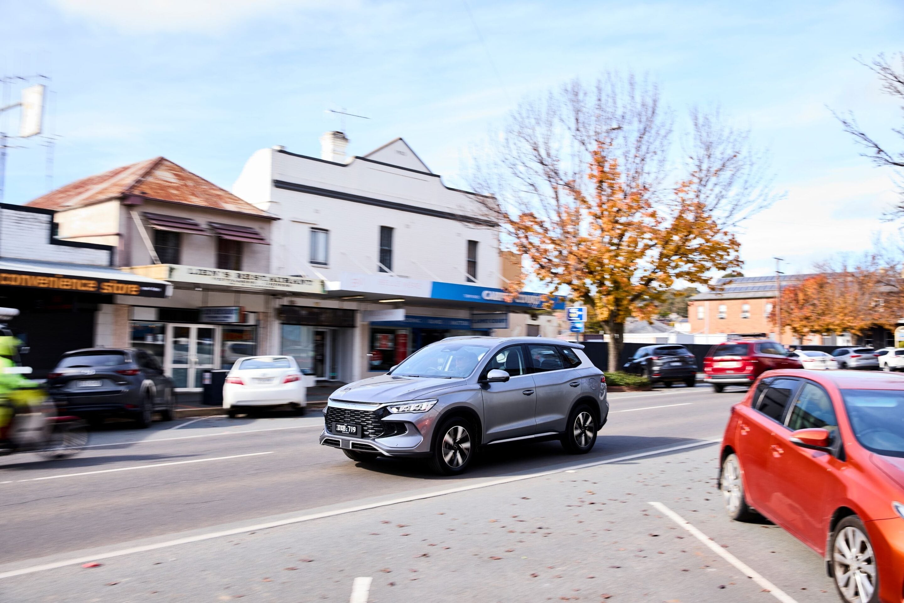 Silver SUV driving on a busy urban street with storefronts and autumn trees blurred in motion behind it, daytime city scene.