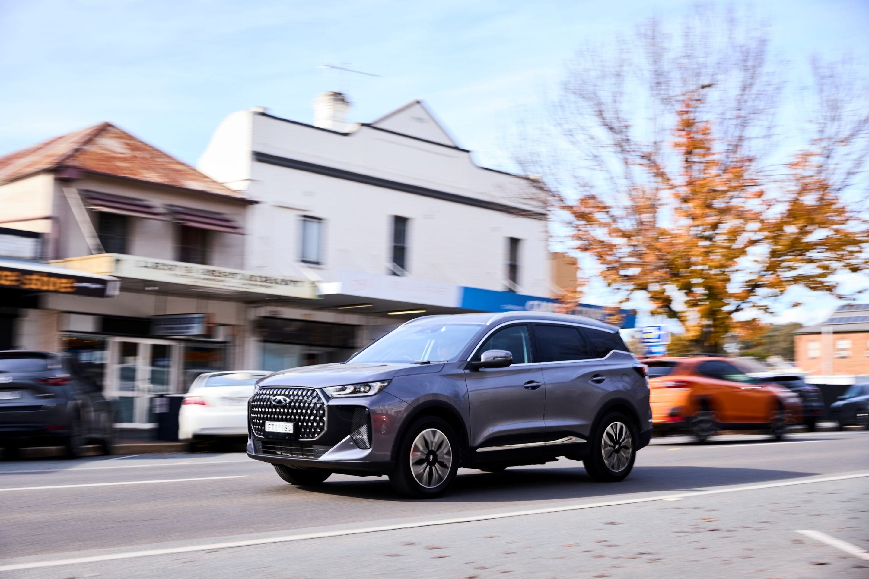 Gray SUV driving on a city street with storefronts and autumn trees in the background, motion blur suggesting speed.