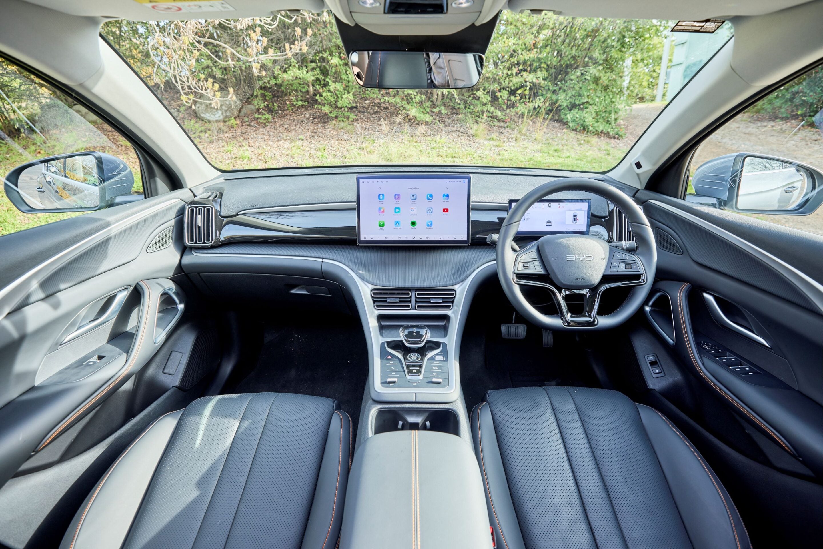 Interior of a modern car cabin featuring a large central tablet-style dashboard screen, a digital instrument display, and black leather seats with orange stitching.