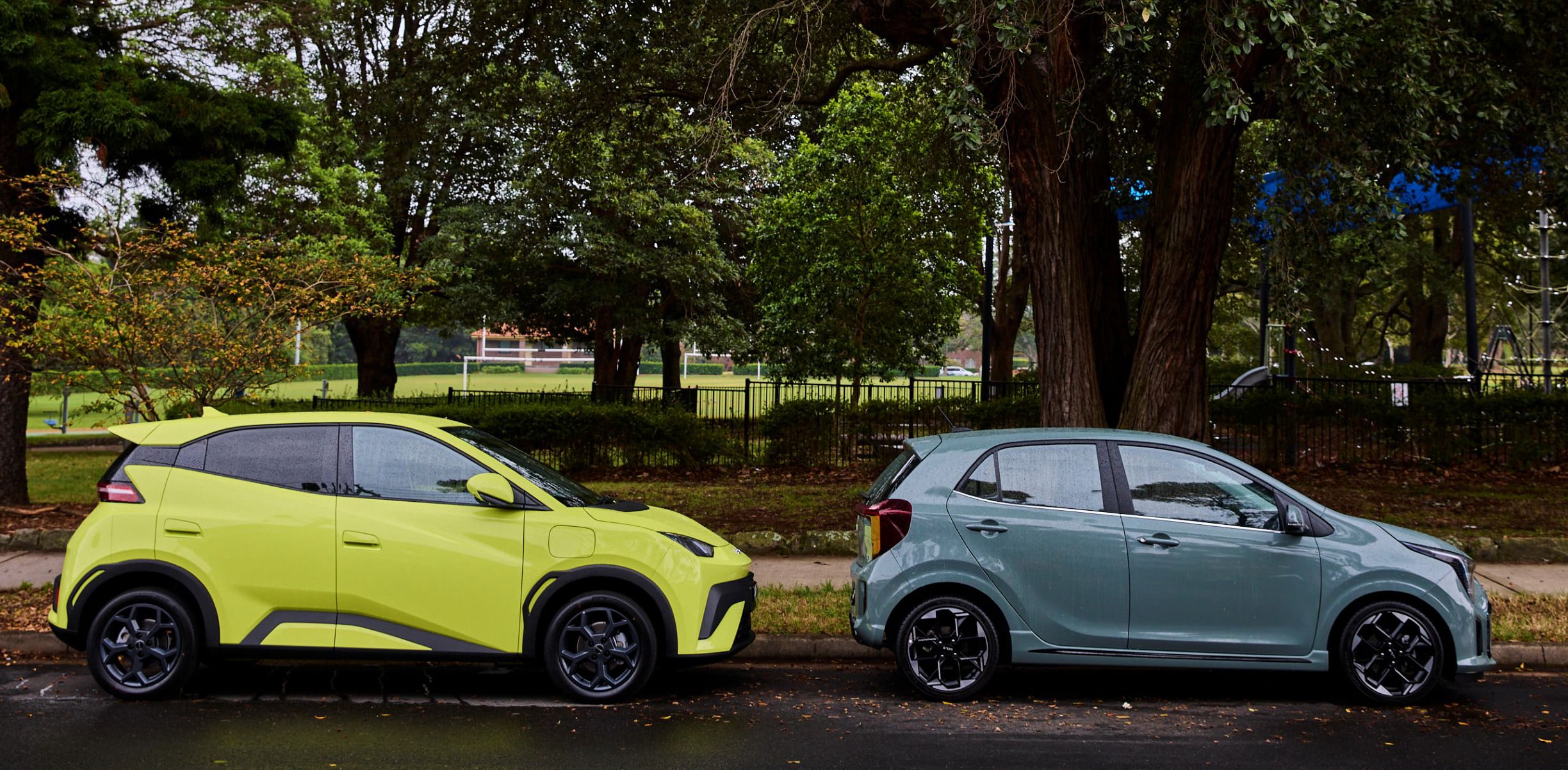 Two modern cars parked side by side in a park-like lot with green trees in the background, the left car bright yellow-green and the right car pale blue-gray.