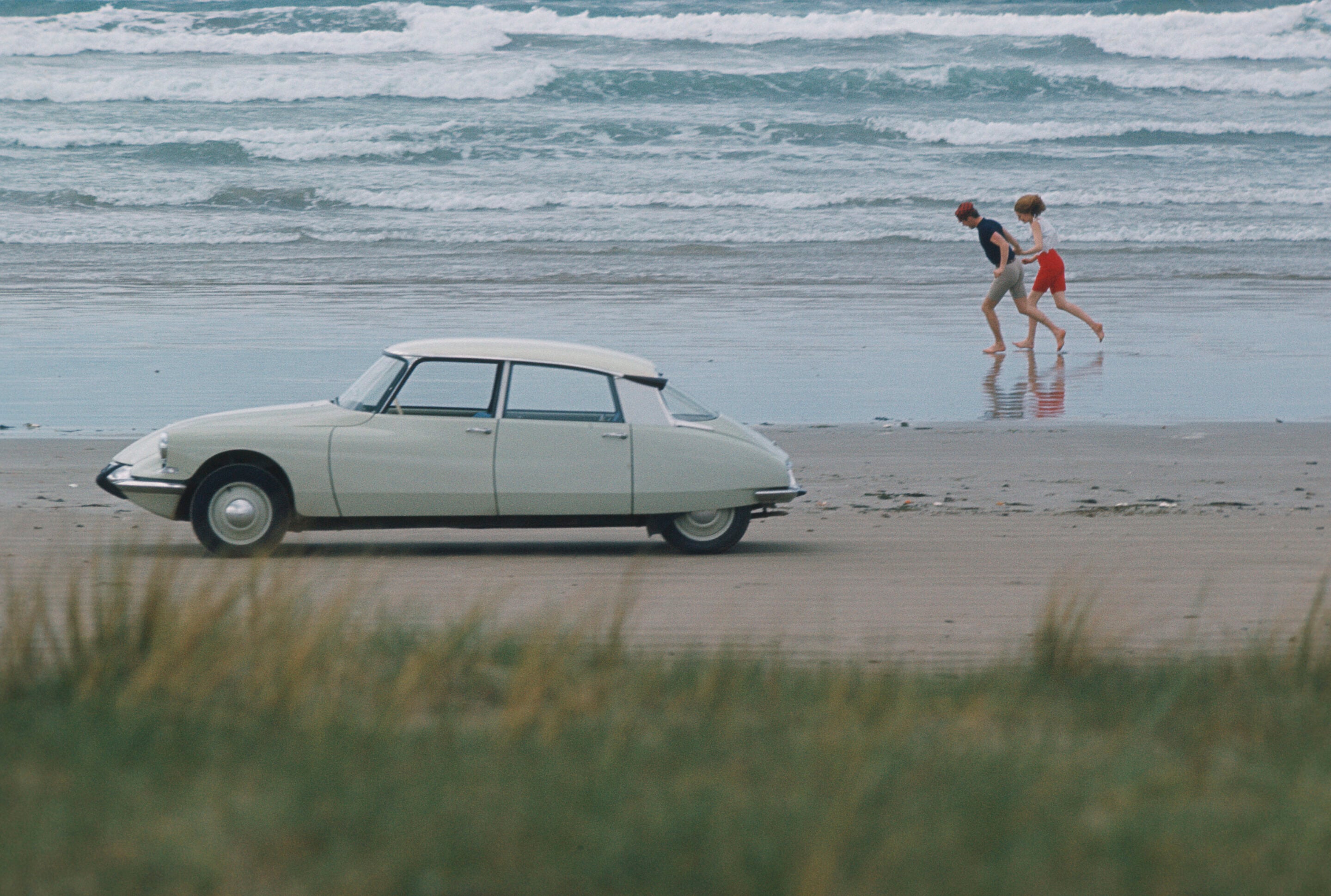 Vintage light-colored car parked on a wet beach while a man and woman walk along the shoreline with waves in the background.