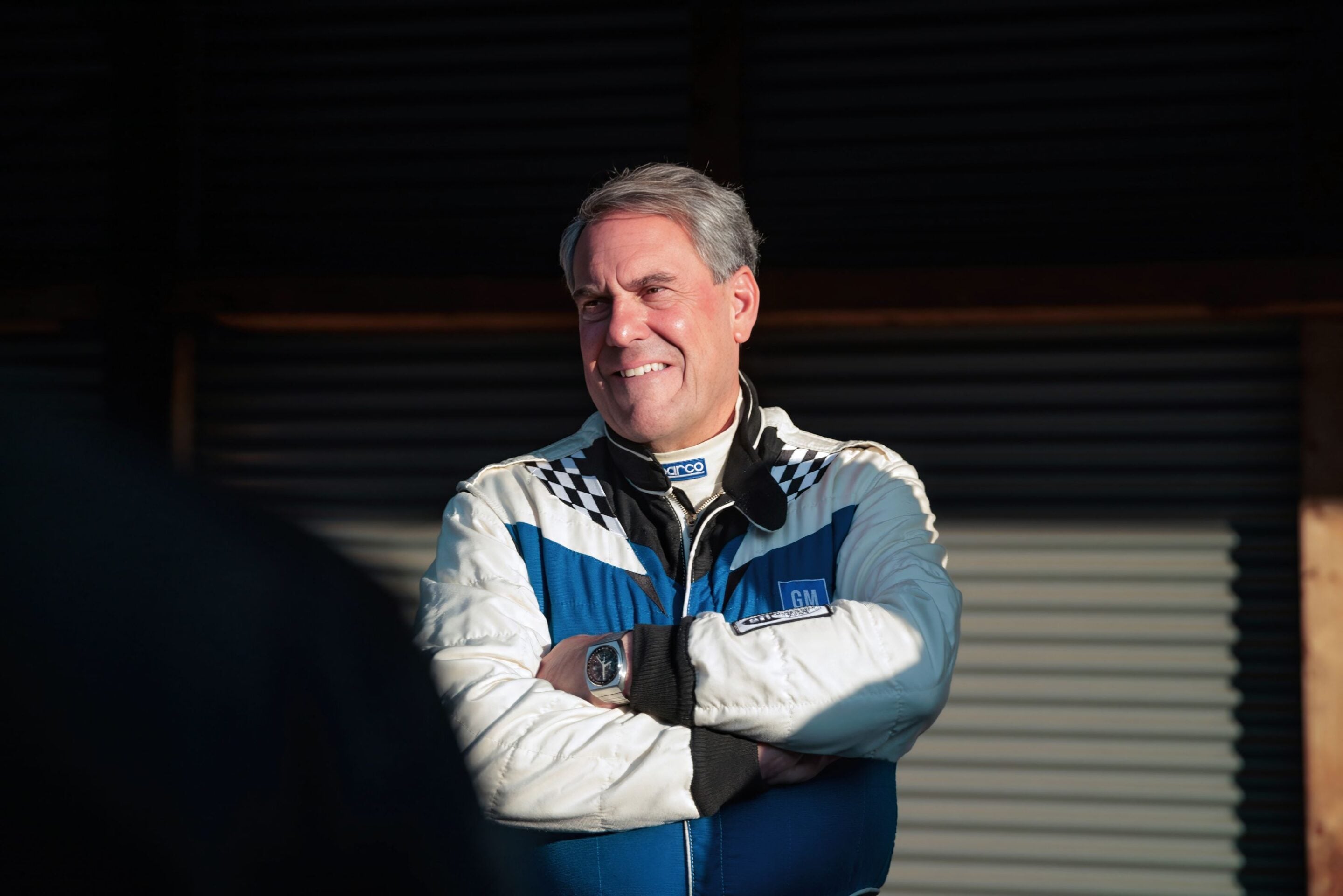 Smiling older man in a blue-and-white racing suit with arms crossed, standing in a dim garage or pit area.