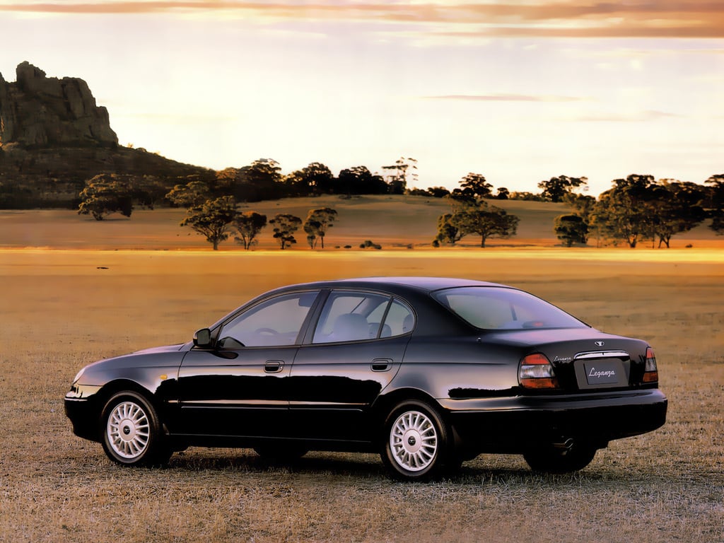 Black sedan parked on dry grass with a distant treeline and rocky hill at sunset, city-less landscape behind it.