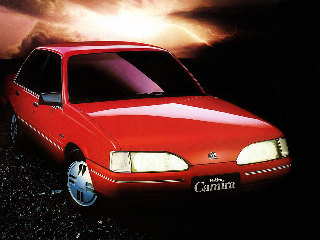 Front-left view of a red Holden Camira car on rocky ground with a dramatic, lightning-filled sky in the background.