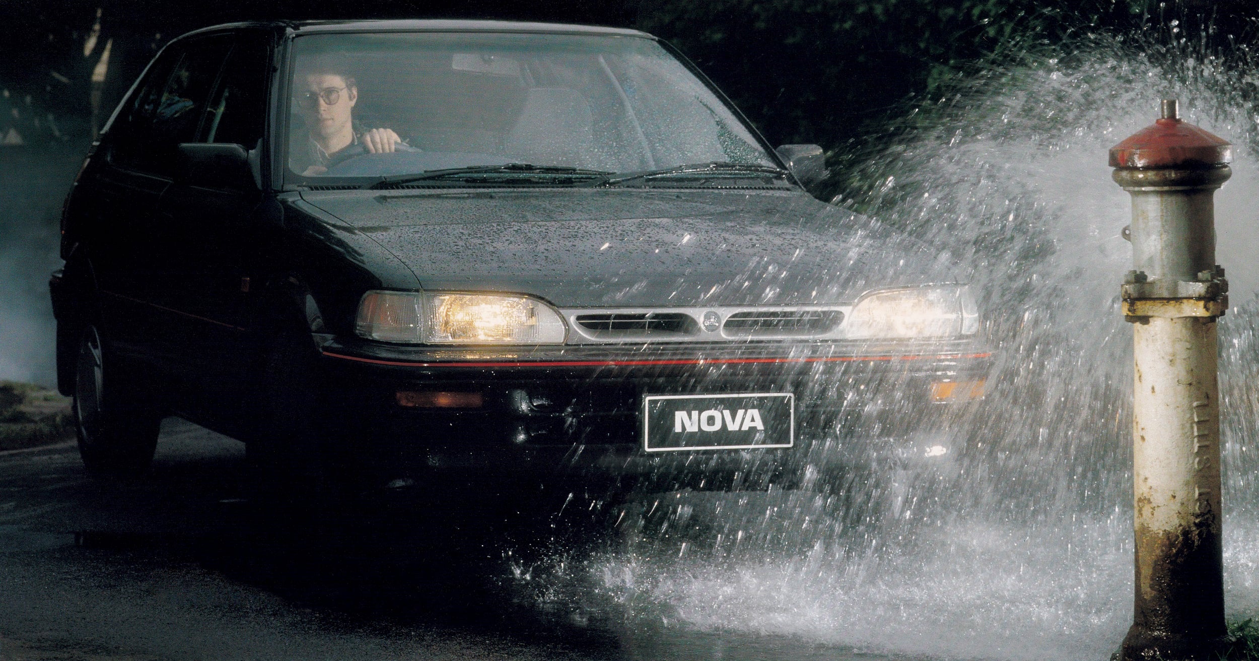 Black sedan drives through a heavy water spray from a fire hydrant, windshield wipers active, NOVA license plate visible behind the spray.
