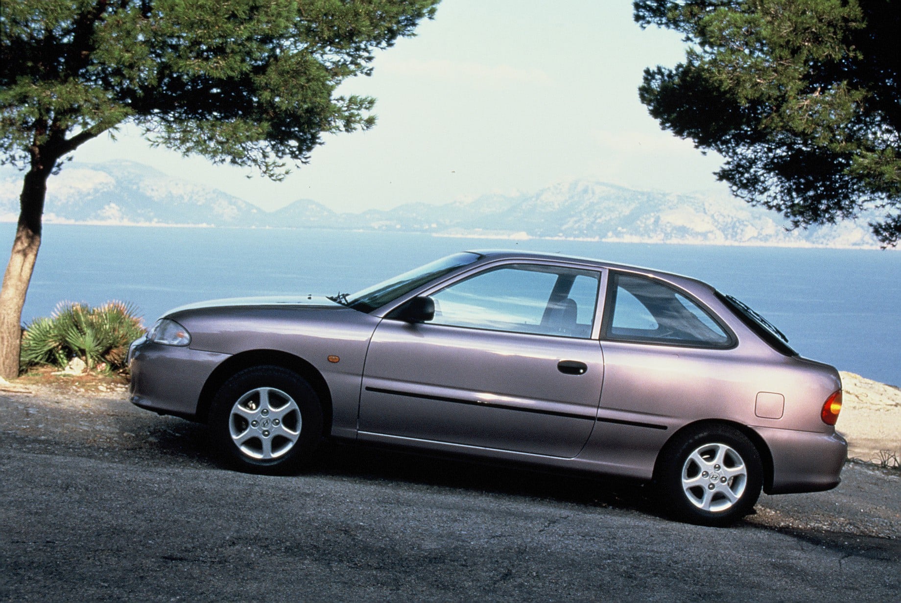 Silver sedan parked on a coastal road with the blue sea and distant mountains framed by trees at the sides.