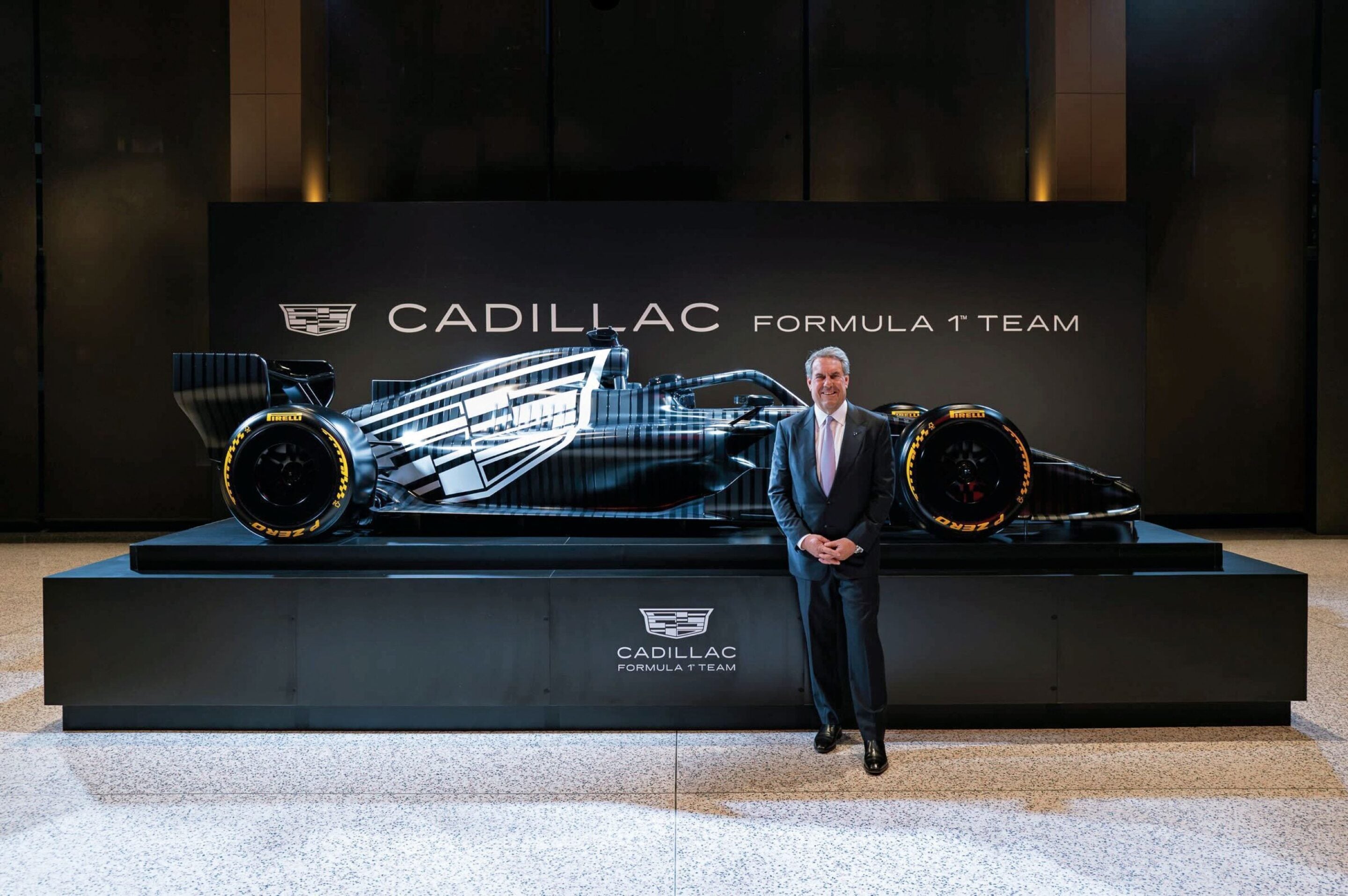 Man in a suit stands beside a blue-and-white Cadillac Formula 1 race car on a dark display platform. (Cadillac Formula 1 Team exhibit)