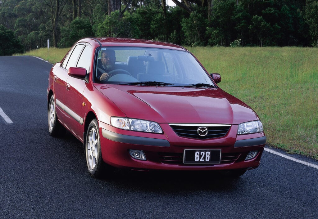 Front-three-quarter view of a red sedan on a paved road with a man visible in the driver's seat and a forested background, license plate reading 626.