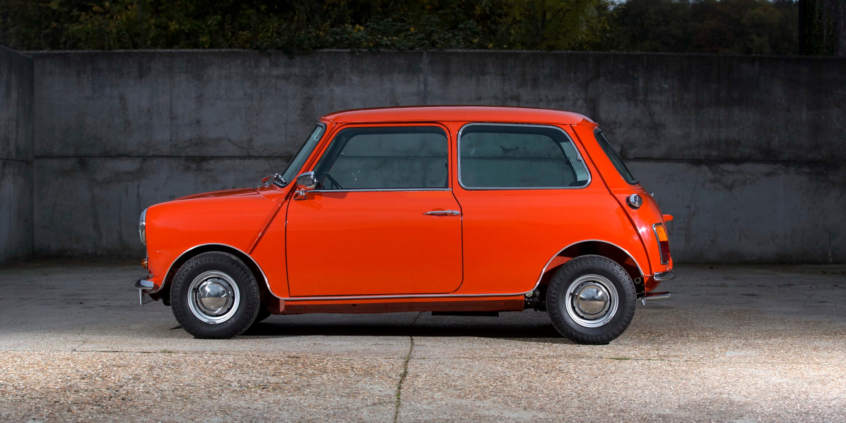 Orange vintage compact car in profile view parked on a concrete surface with a gray wall in the background, showcasing classic round headlights and chrome trim.