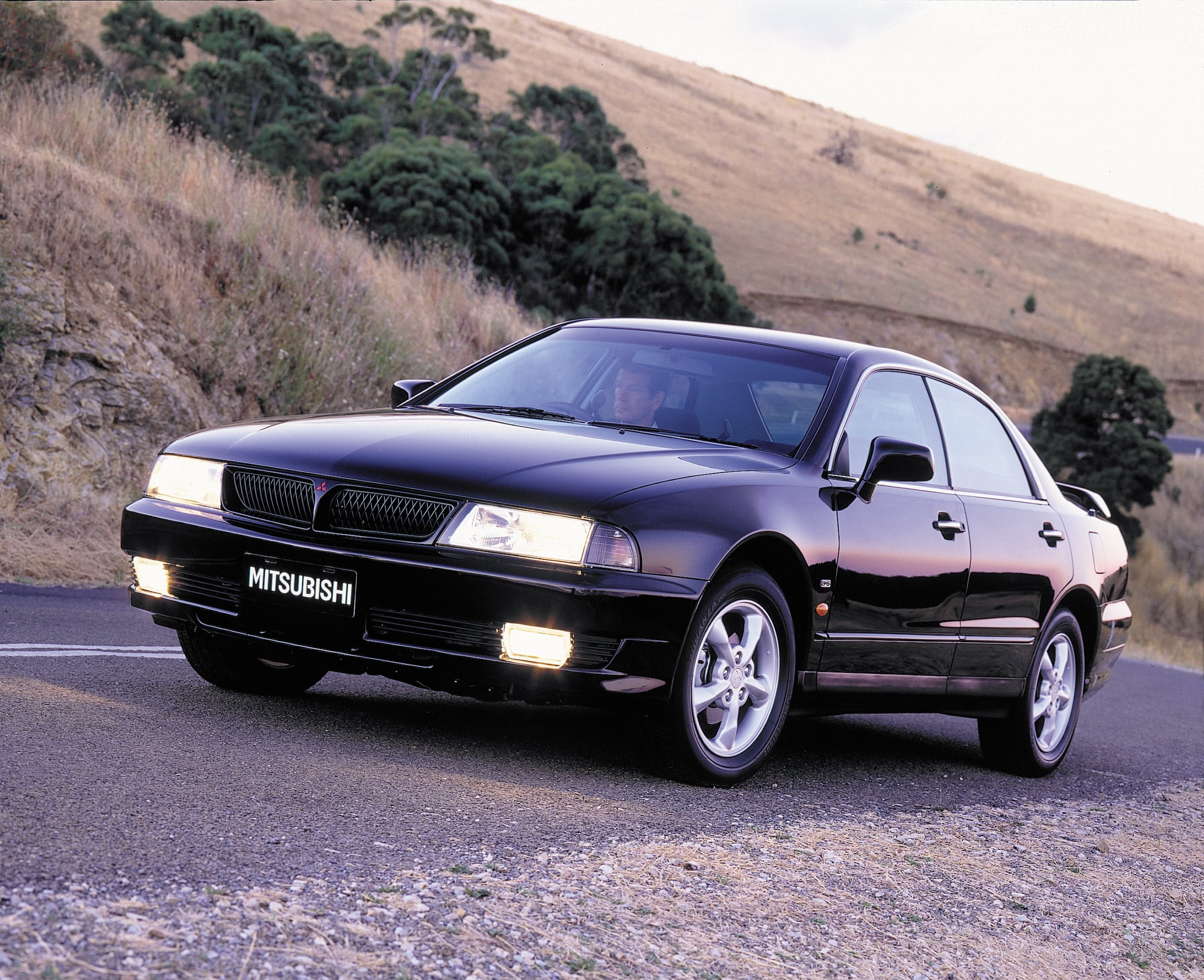 Black Mitsubishi sedan parked or driving on a winding hillside road, front angled toward the camera with a driver visible inside.