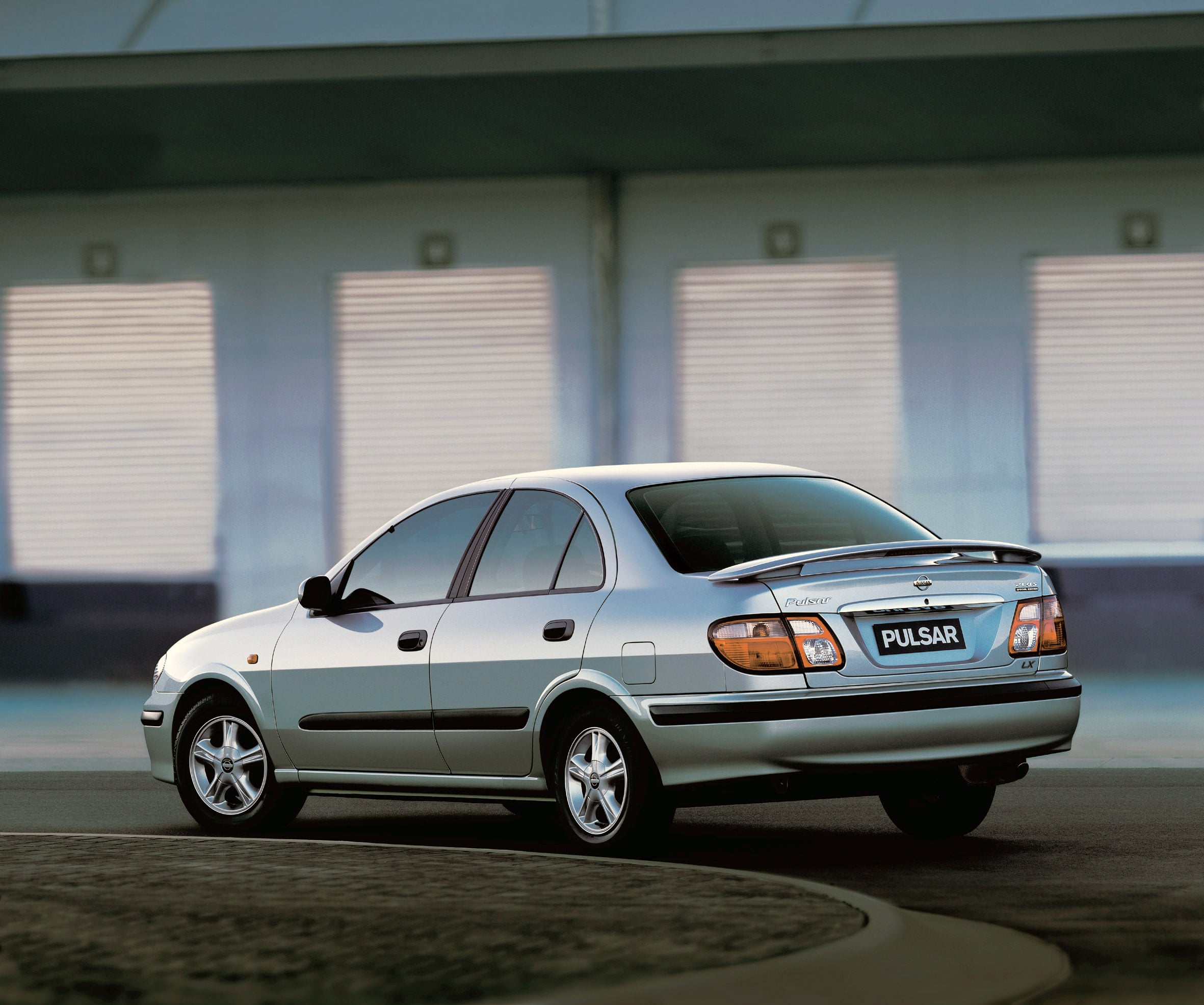 Rear three-quarter view of a light blue Nissan Pulsar sedan parked in a lot near a building with shutters on the background, daytime.