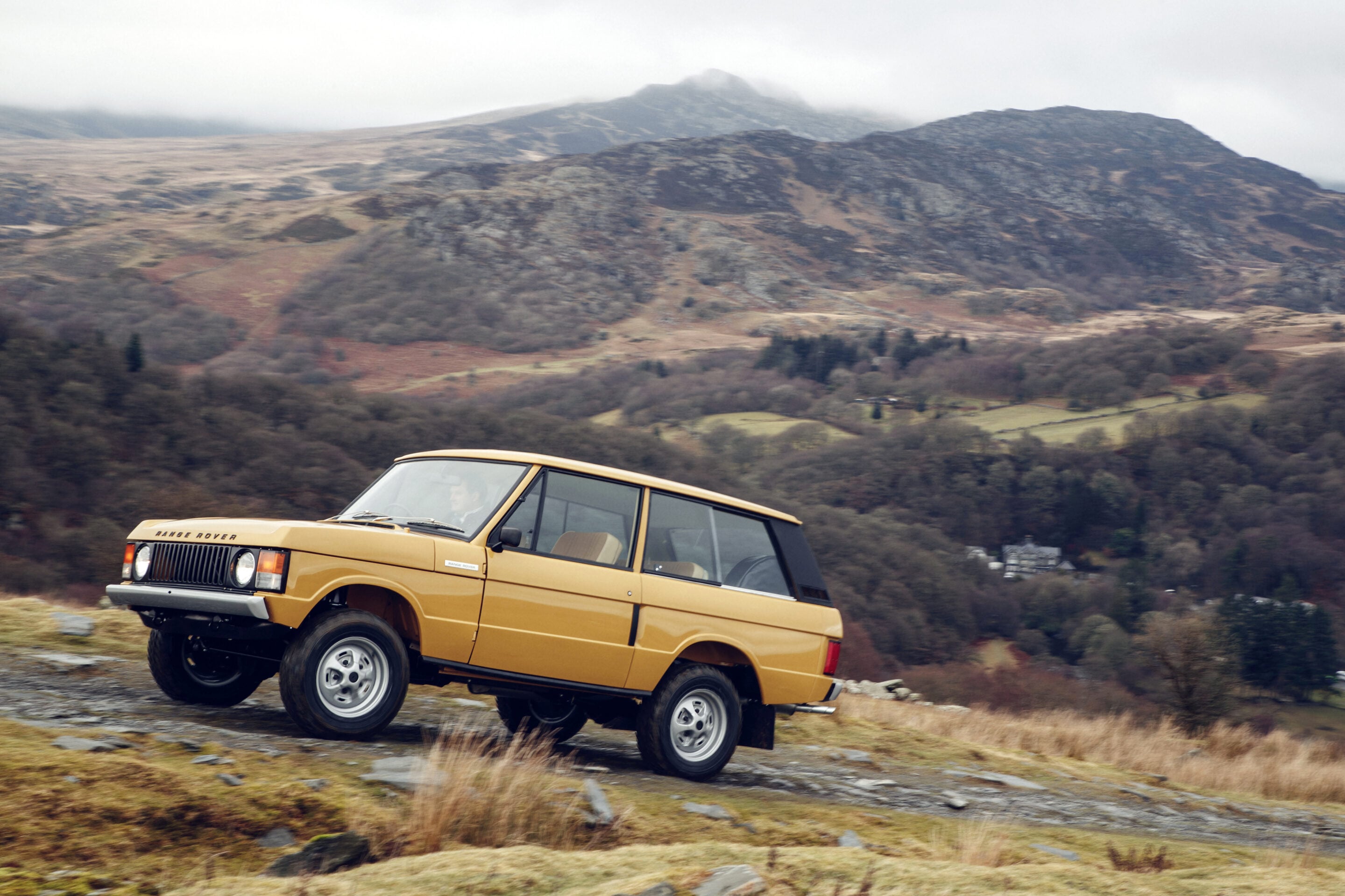 Beige Range Rover driving on rough, rocky terrain with a rugged mountain landscape in the background under a cloudy sky.