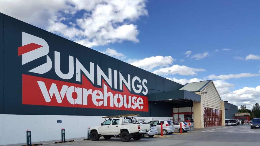 Storefront of Bunnings Warehouse with large white and red logo on a dark blue wall, and a parking lot with trucks and cars under a blue sky.