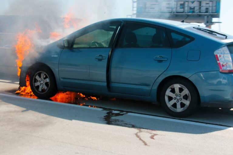 Blue car on fire with orange flames around the front left wheel and rising smoke on a road surface