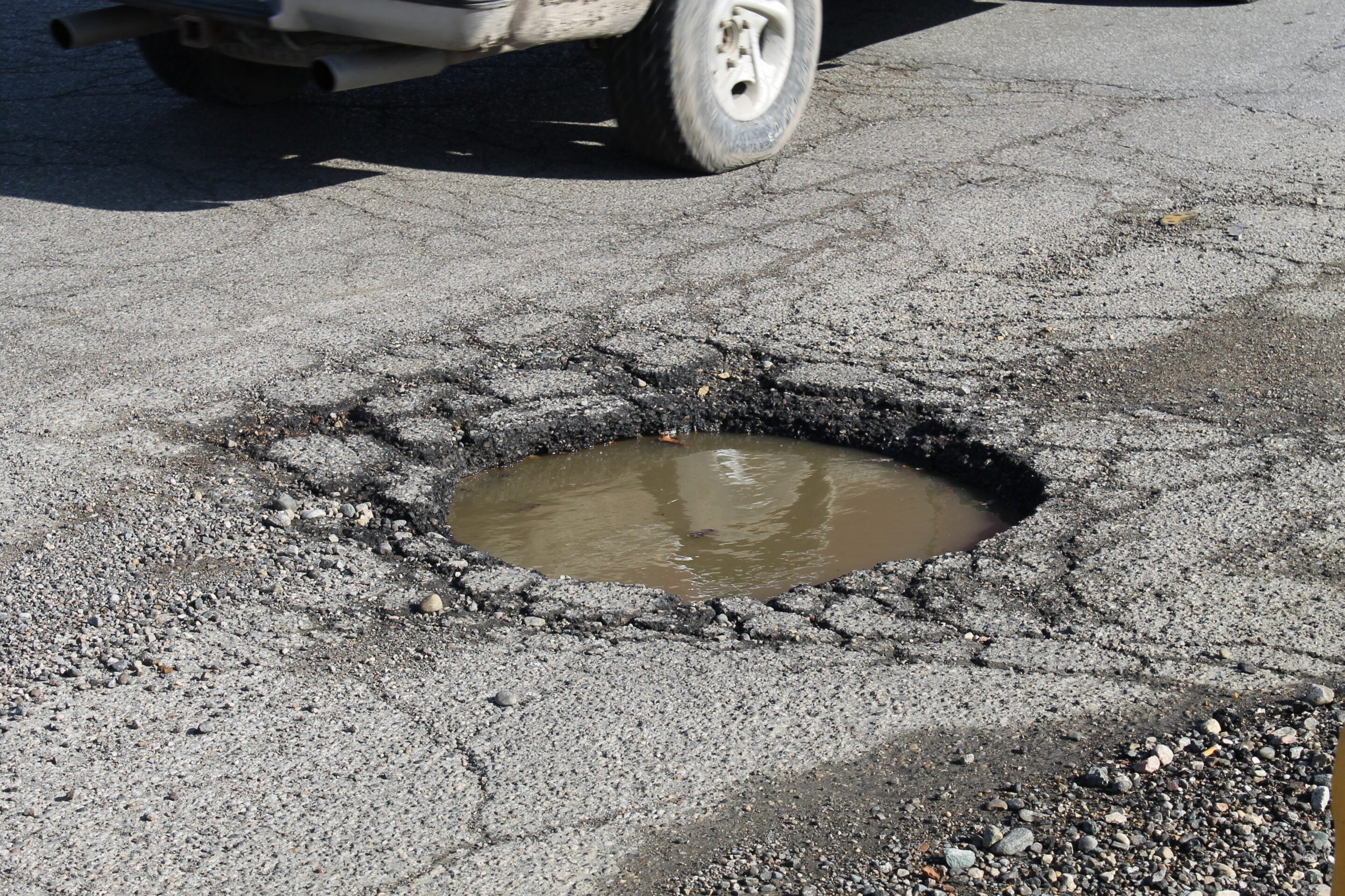 Rear half of a car with exhaust pipes visible, parked beside a large pothole filled with muddy water in a cracked asphalt road surface.