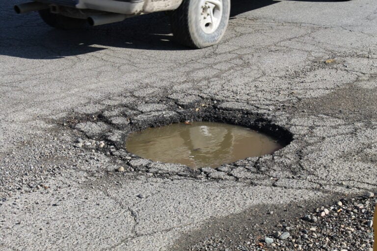Rear half of a car with exhaust pipes visible, parked beside a large pothole filled with muddy water in a cracked asphalt road surface.