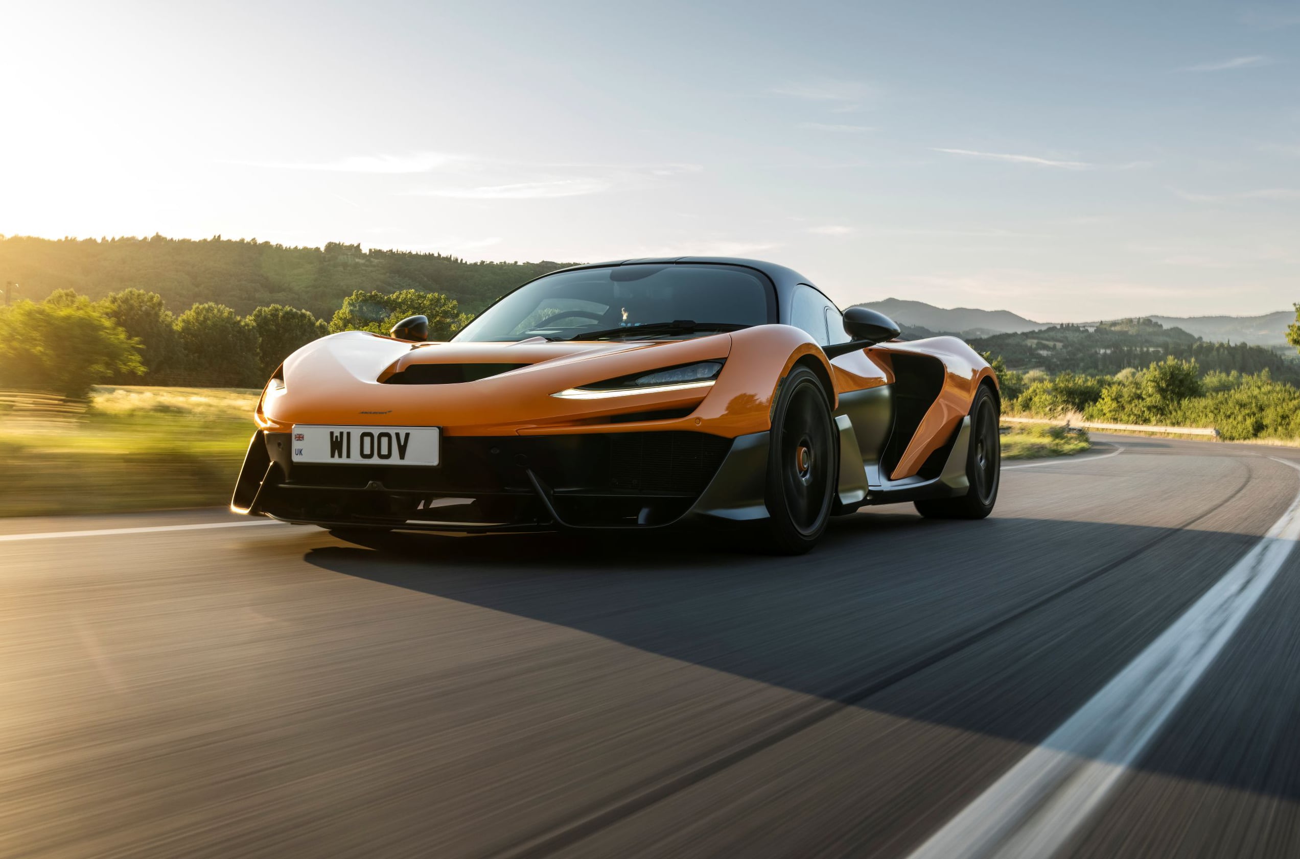 Orange sports car speeding on a curved highway with motion blur and a scenic hillside backdrop at sunset