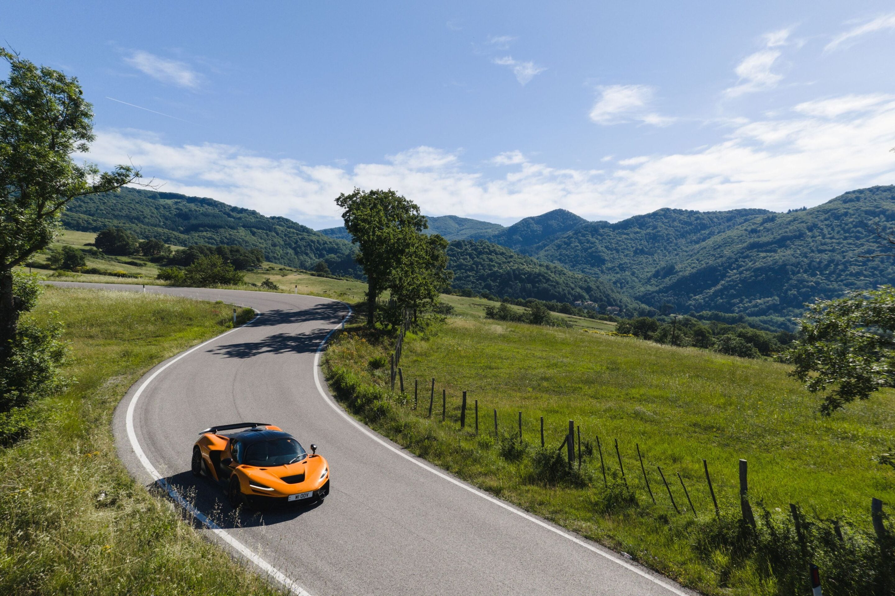 Orange sports car on a winding mountain road with green hills and blue sky.