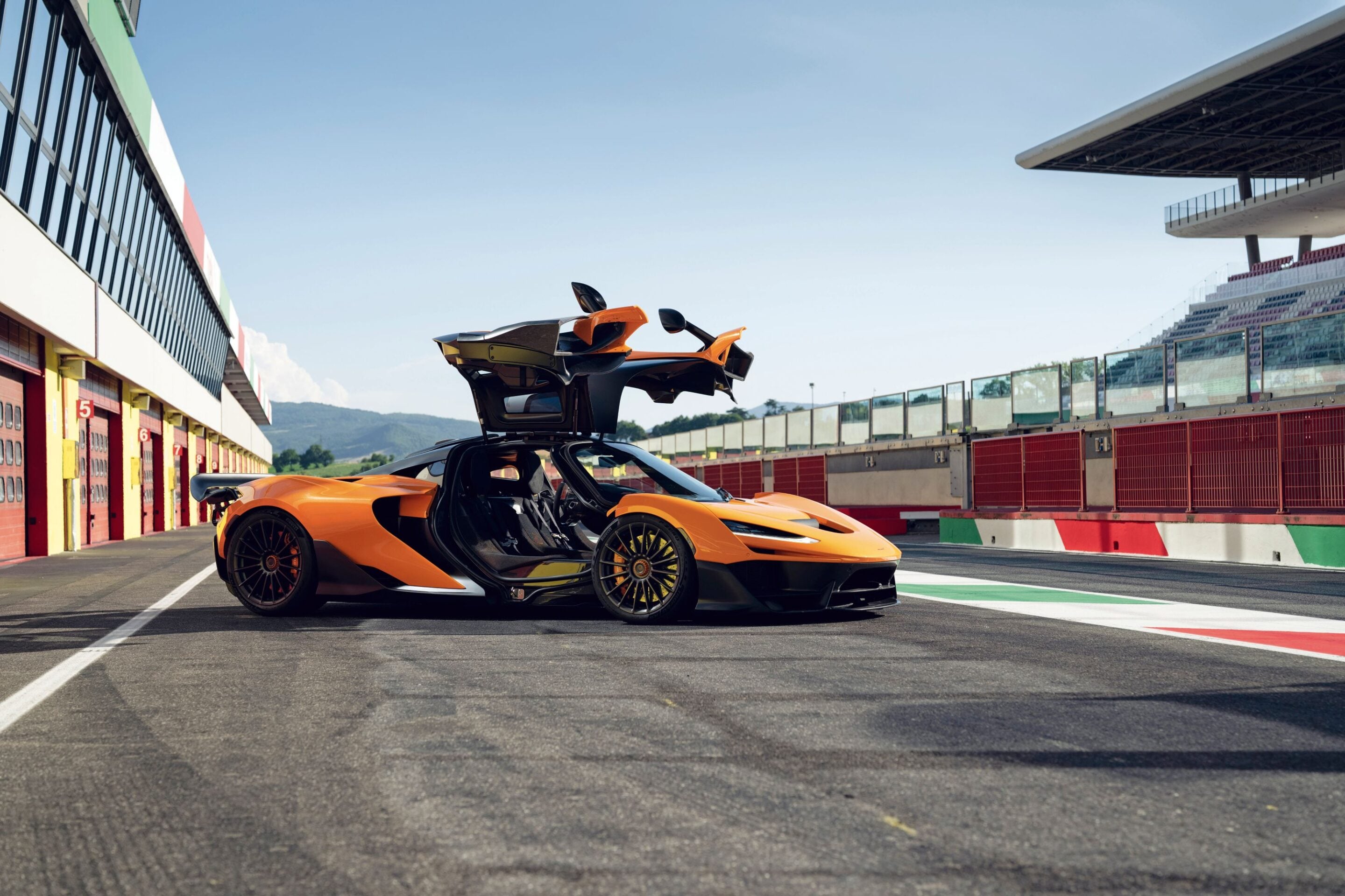 Orange exotic sports car with doors up, parked in pit lane beside red garages at a race track.