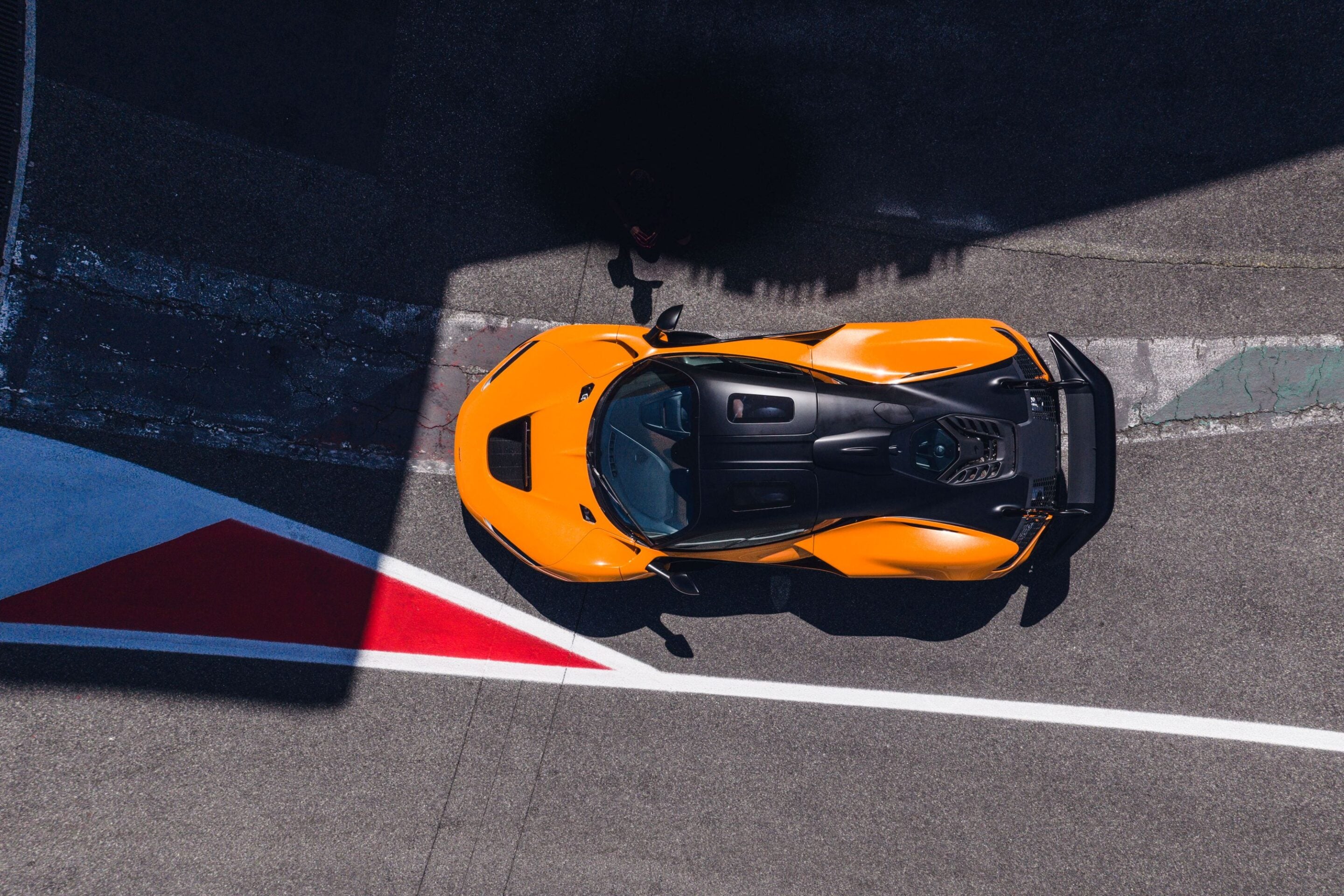 Top-down view of an orange sports car parked on asphalt with white and red track markings nearby.