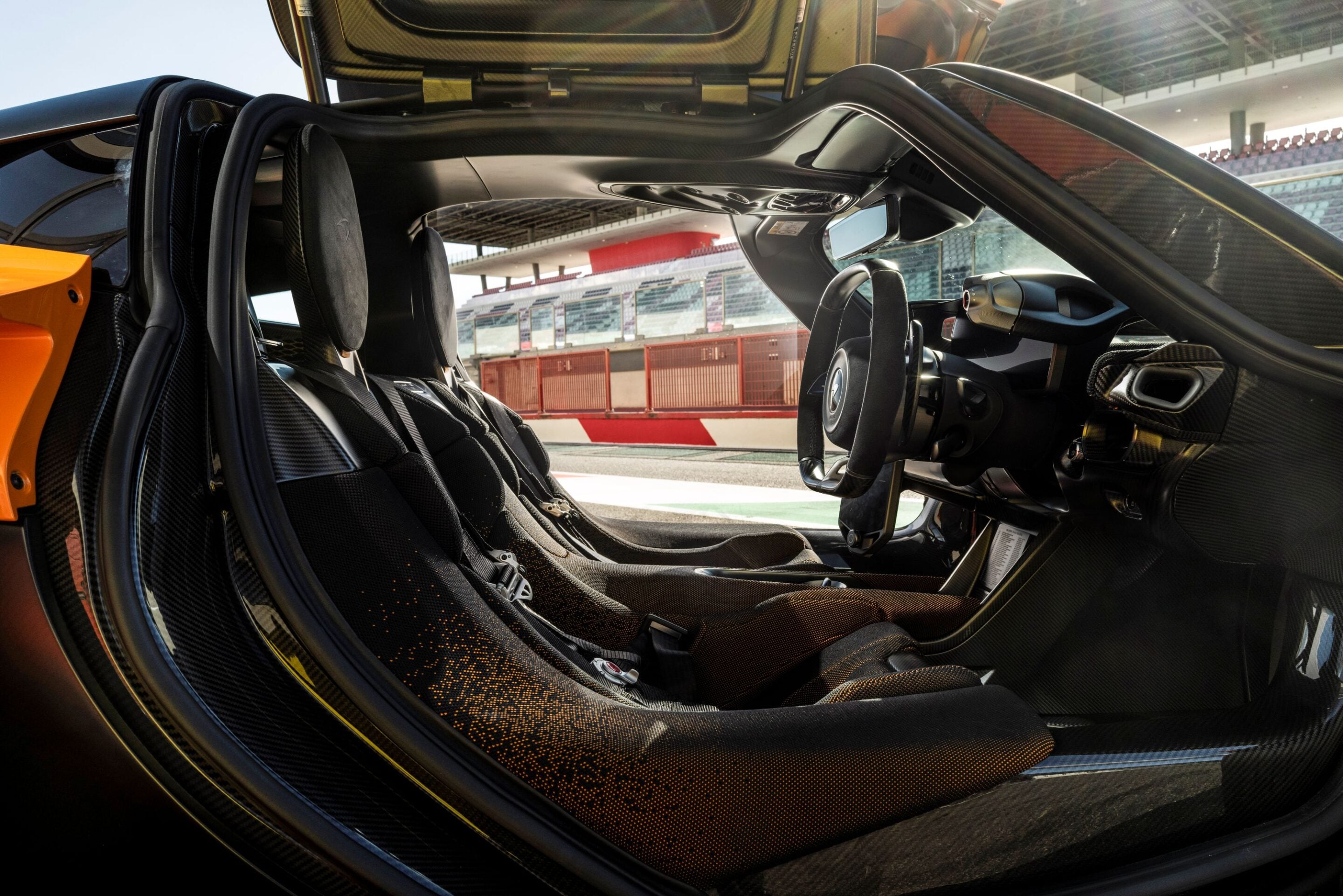 Open gull-wing door reveals a racing-car interior with carbon fiber trim and bucket seats, track visible outside.