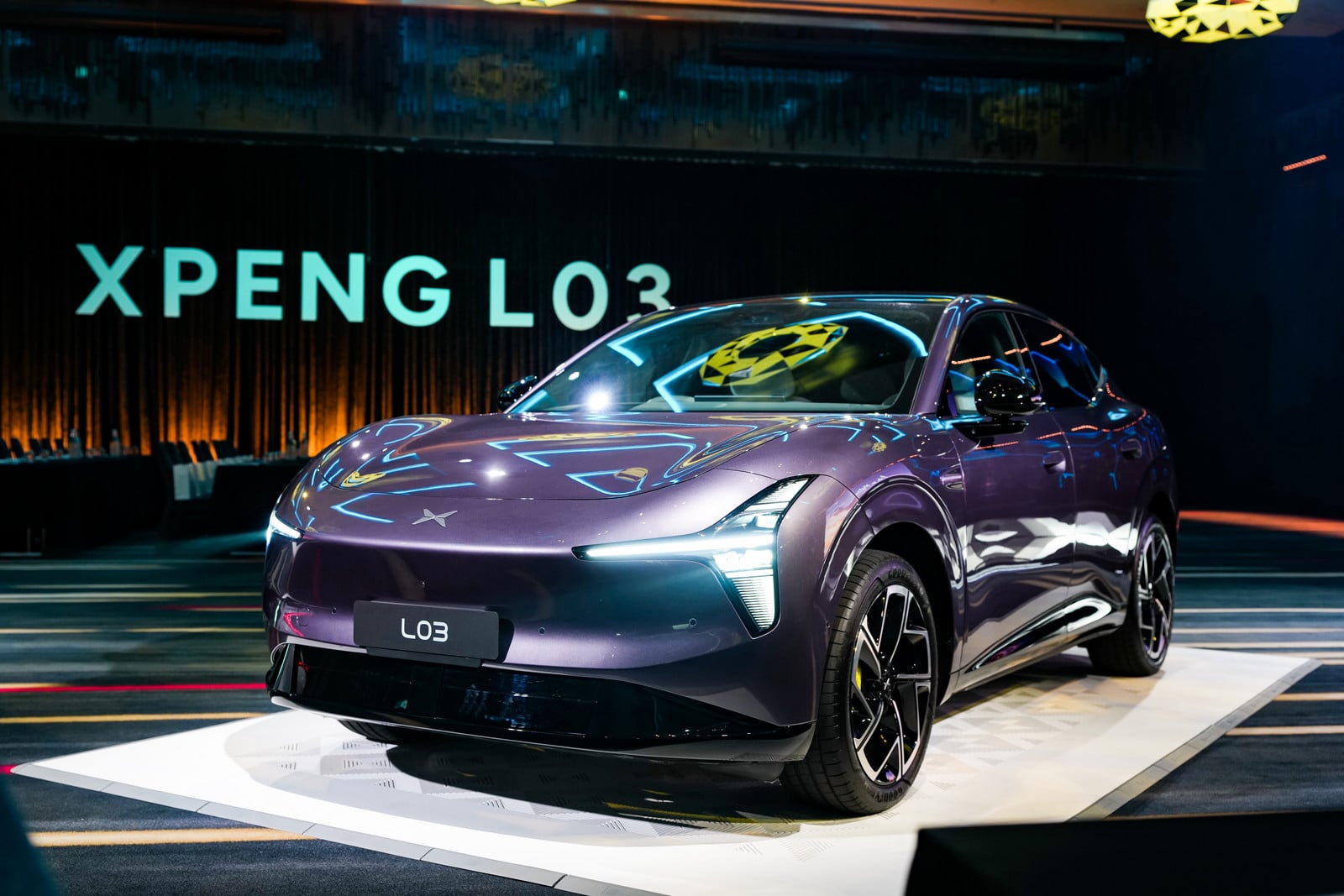 Purple XPENG L03 on a white display platform at an auto show, with neon reflections and a bold backdrop reading XPENG L03.