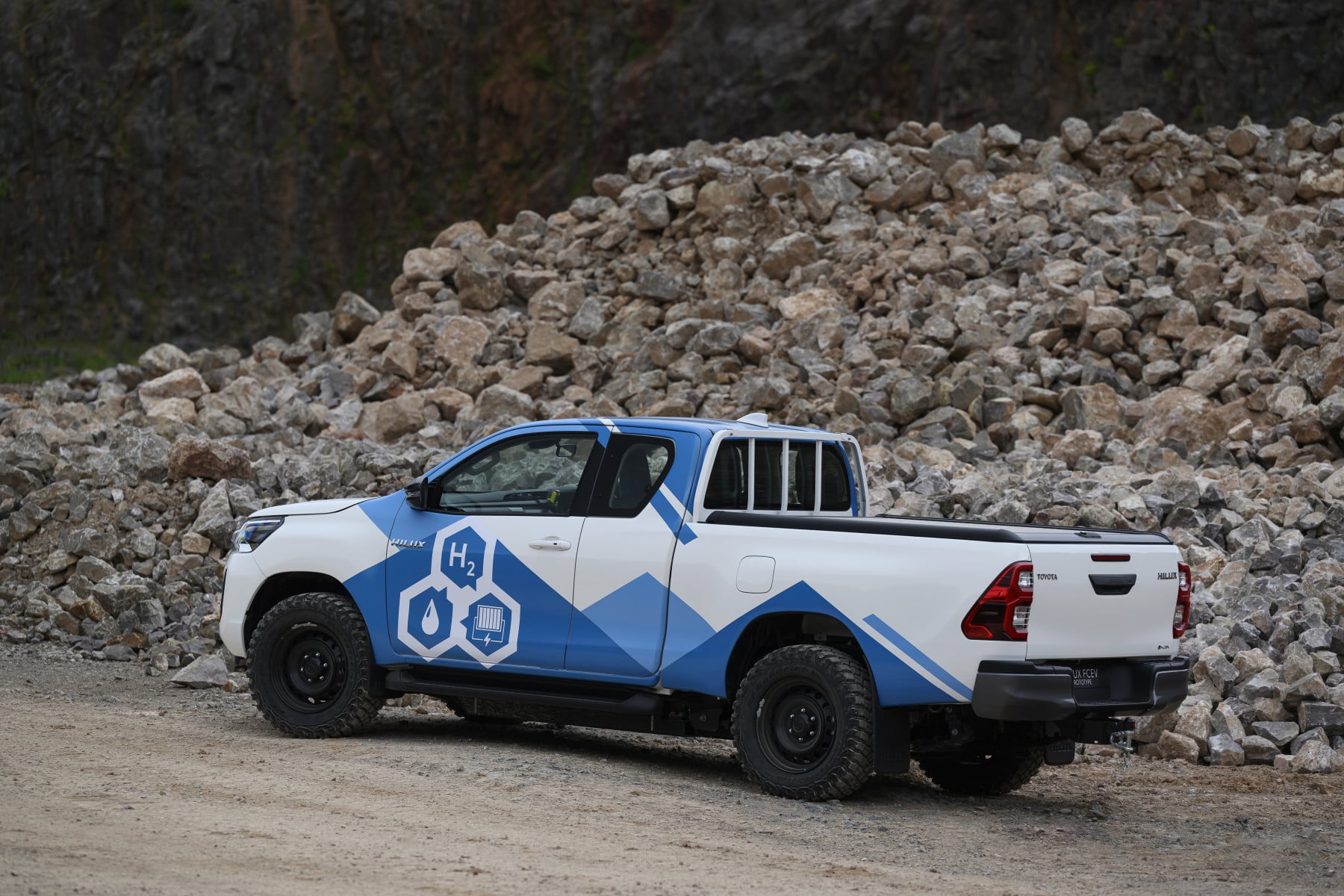 Hydrogen-themed white and blue Toyota Hilux pickup parked beside a rocky quarry pile of stones.