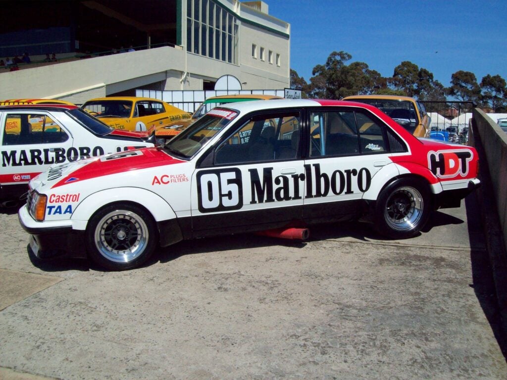 White and red race car with large Marlboro lettering parked in a row of colorful race cars, near a modern building under a clear blue sky, likely at a track paddock.