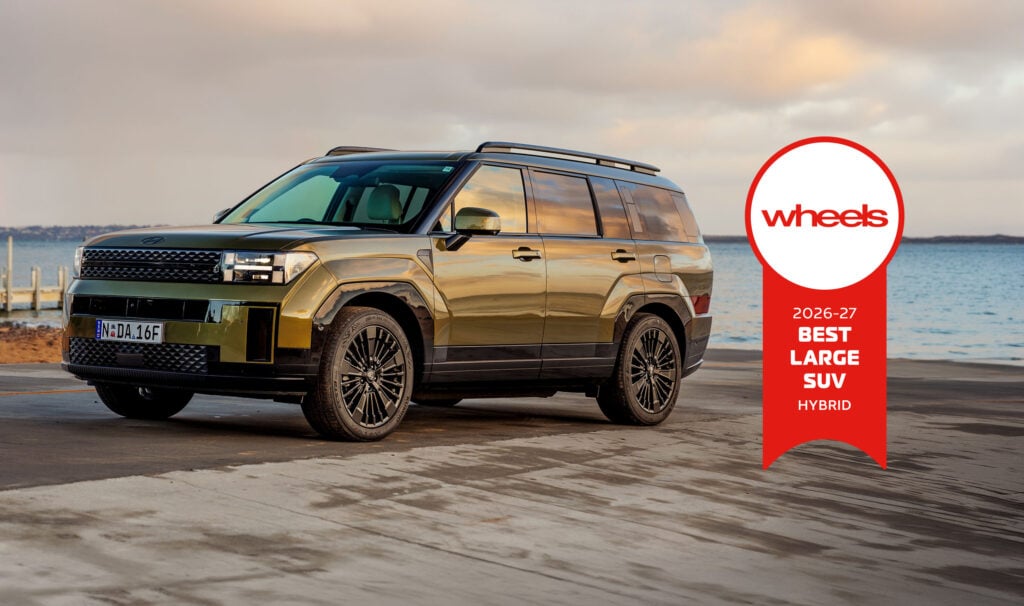 Golden SUV parked by the sea with a red Wheels award badge reading 2026–27 Best Large SUV Hybrid in the foreground.