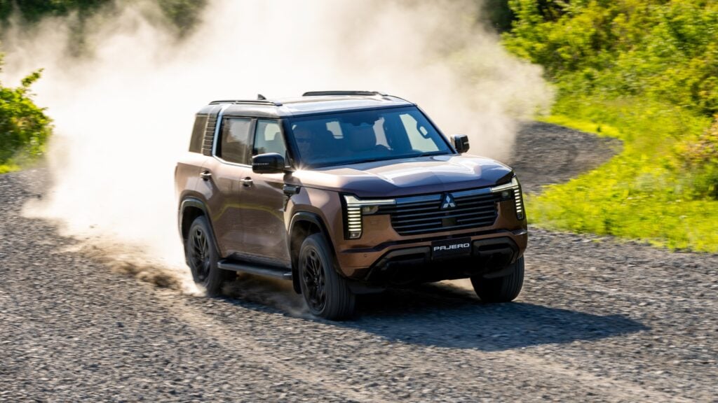 Brown SUV (Pajero) speeding along a gravel road, dust plumes rising behind the tires with green vegetation on the sides.