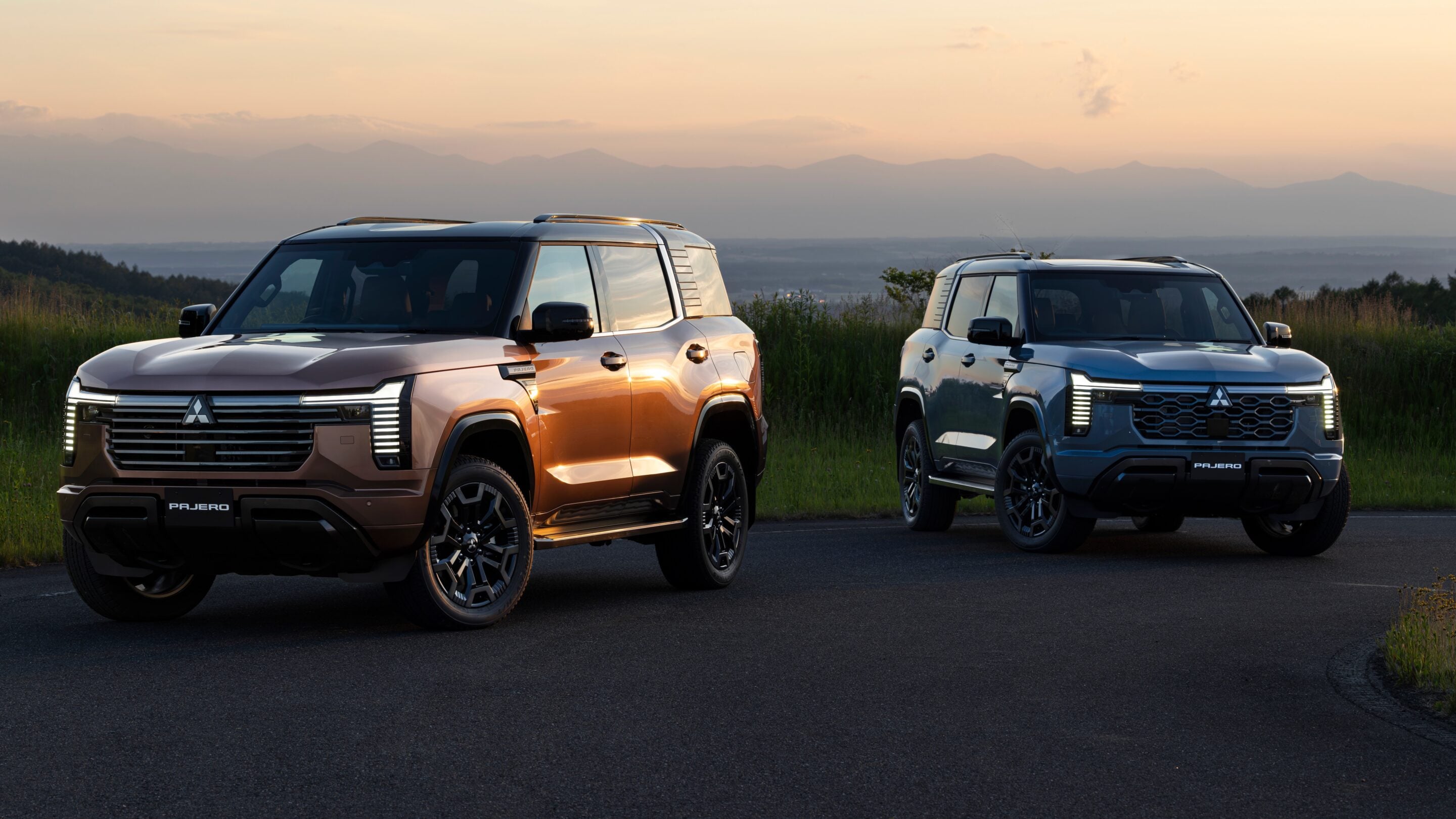Two Mitsubishi Pajero SUVs parked on a paved road at sunset, with a distant mountain range and tall grass in the foreground.