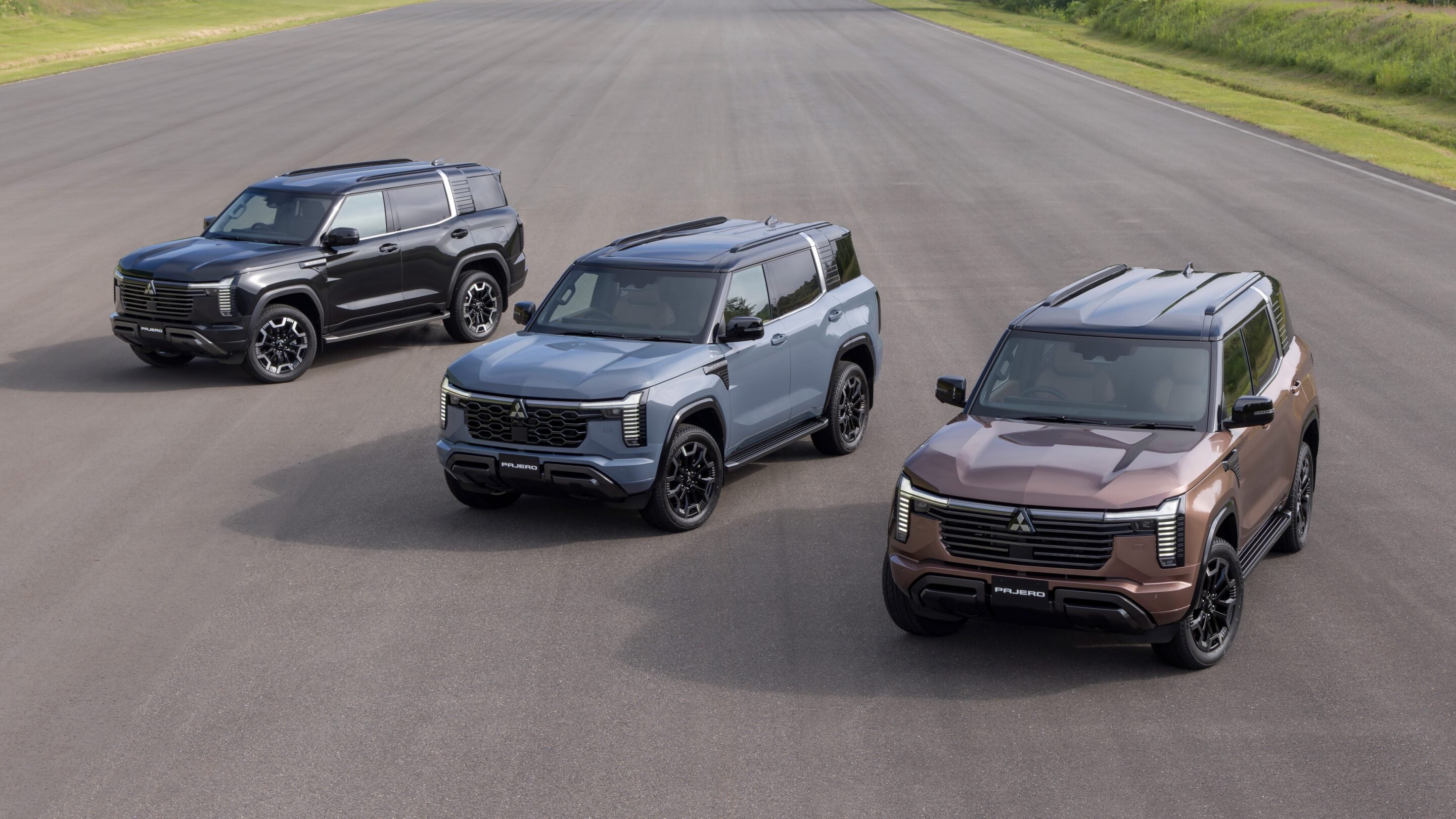 Four Mitsubishi Pajero SUVs in gray, blue, and brown parked on an empty race track, viewed from above with prominent grilles and LED headlights on display.