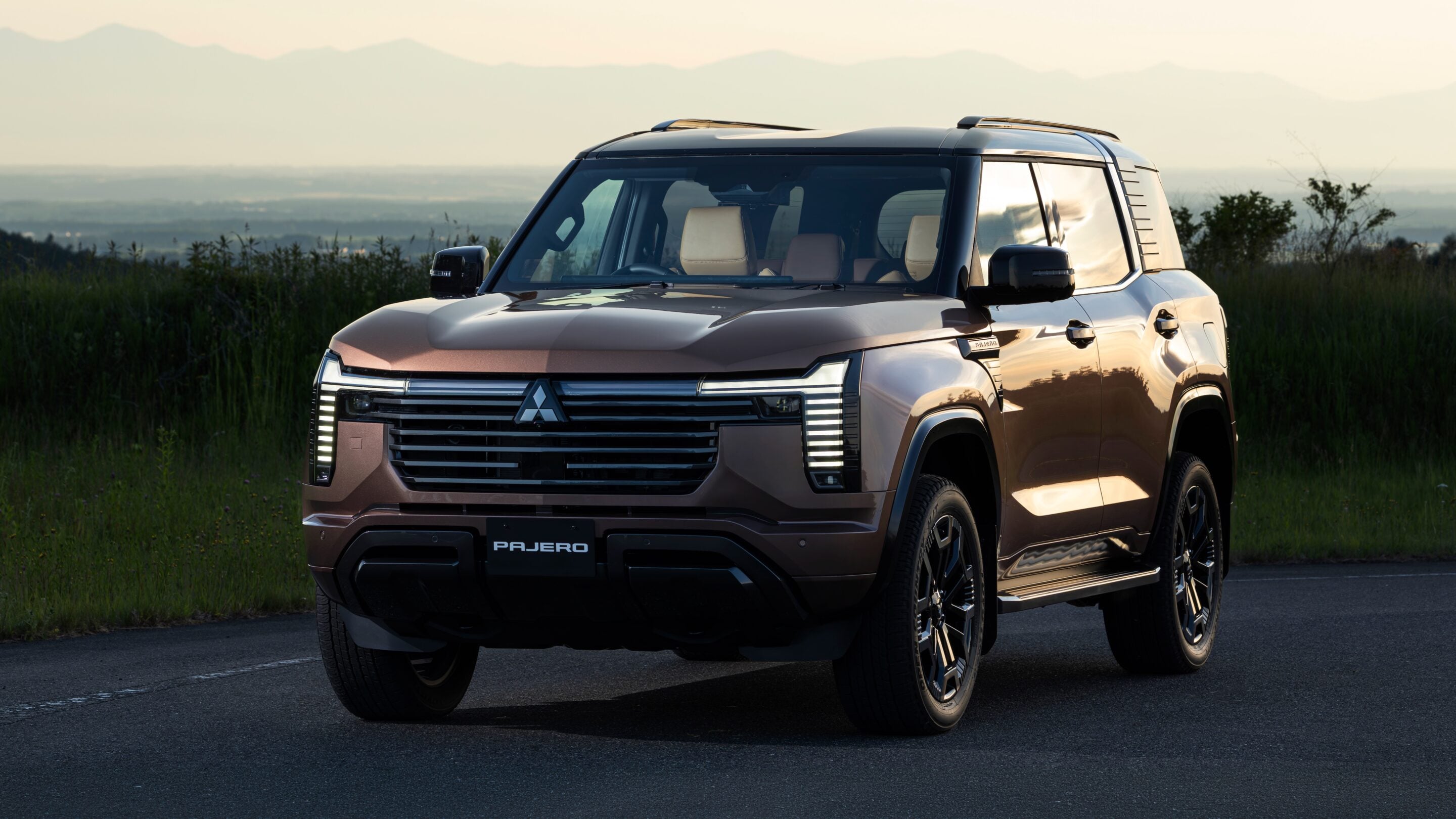 Front three-quarter view of a bronze Mitsubishi Pajero SUV parked on a paved lot with a grassy field and distant mountains in the background, during sunset light.