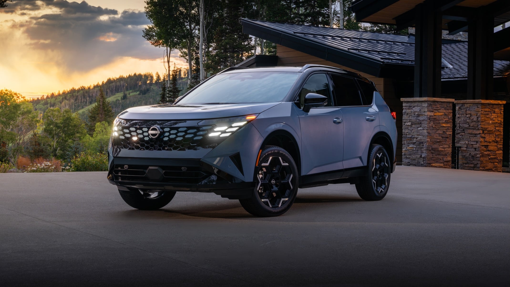 Gray Nissan SUV parked in a driveway beside a modern house with stone columns at sunset, hills in the background.